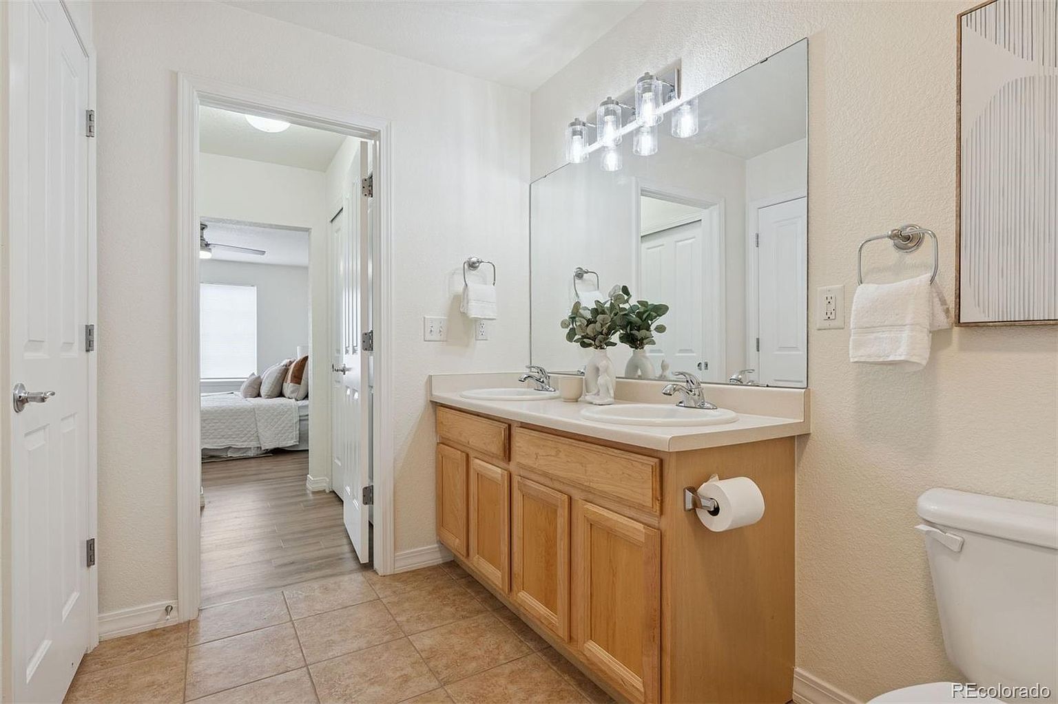 This is a well-lit primary bathroom featuring a double vanity with light wood cabinetry and white countertops. A large mirror hangs above the sinks, reflecting the bright lighting and the adjacent bedroom. The bathroom has neutral-toned tile flooring and a clean, inviting aesthetic.