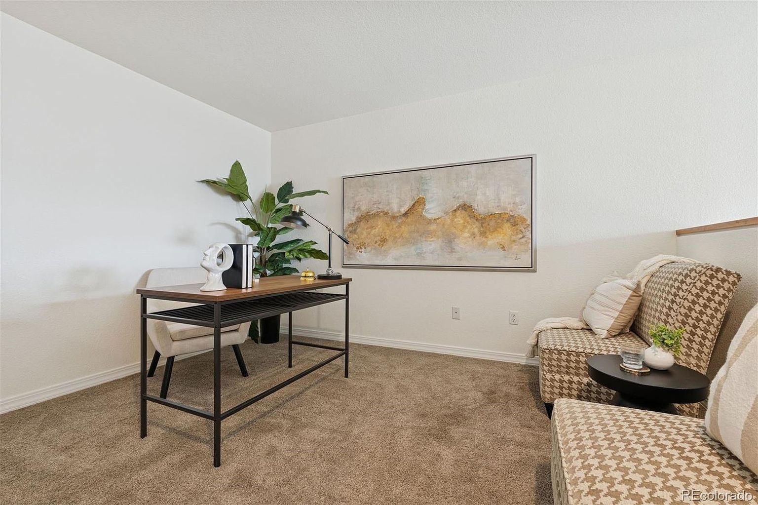 This is an interior shot of a home office or study. The room features a desk with a plant and decorative items, a framed piece of art on the wall, and a patterned armchair with a small side table. The space is carpeted and has white walls, creating a bright and inviting atmosphere.