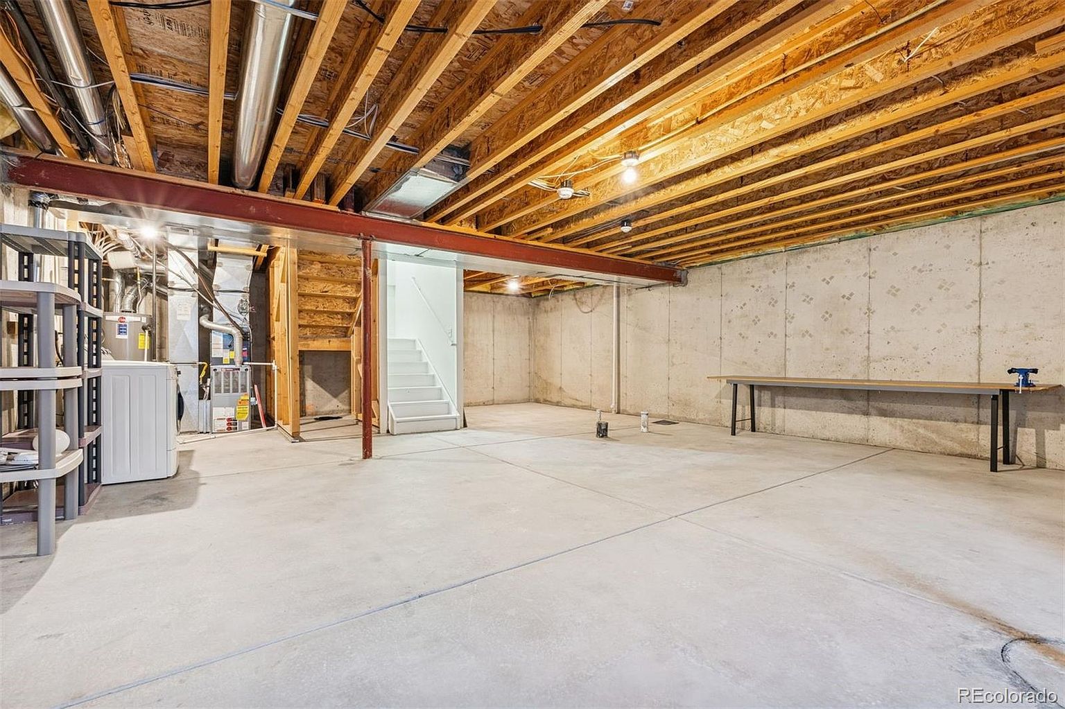 This is a spacious unfinished basement featuring exposed wooden ceiling beams and a concrete floor. A red steel beam runs across the ceiling, and there are visible pipes and ductwork. The basement includes stairs leading to another level and a workbench along one wall, offering potential for customization and additional living space.