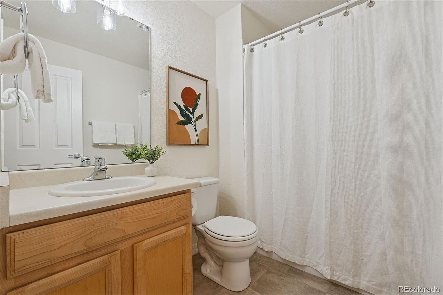 This is a well-lit bathroom featuring a light wood vanity with a white countertop and sink. A framed art piece hangs above the toilet, and a white shower curtain covers the bathtub. The overall impression is clean and functional, suitable for a guest bathroom.