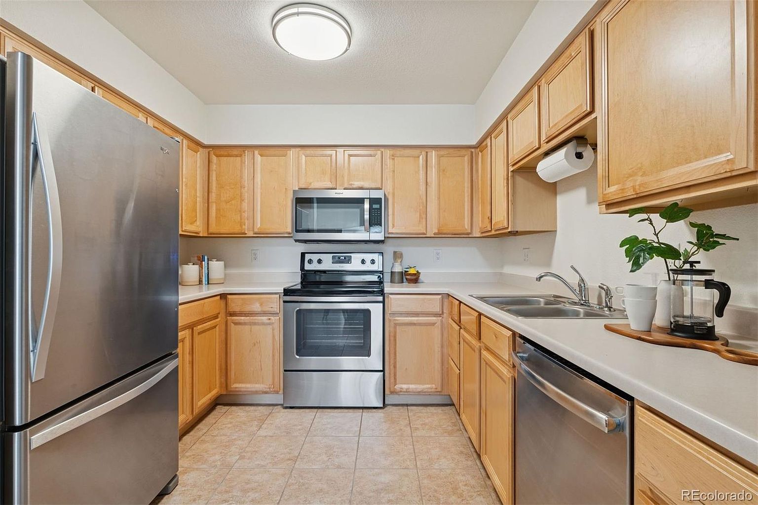 This is a well-lit kitchen featuring light wood cabinetry, stainless steel appliances, and light countertops. The kitchen has a U-shaped layout with a stainless steel refrigerator on the left, a stainless steel oven and microwave in the center, and a sink and dishwasher on the right. The flooring is tiled, and the overall impression is clean and functional.