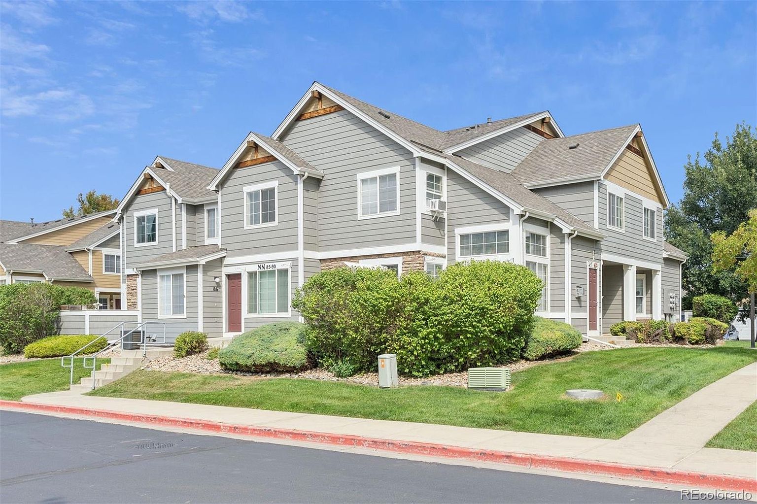 This is a front view of a well-maintained townhouse complex with a gray exterior and white trim. The building features multiple gabled roofs and stone accents, adding visual interest. Lush green landscaping surrounds the property, enhancing its curb appeal and creating a welcoming atmosphere.