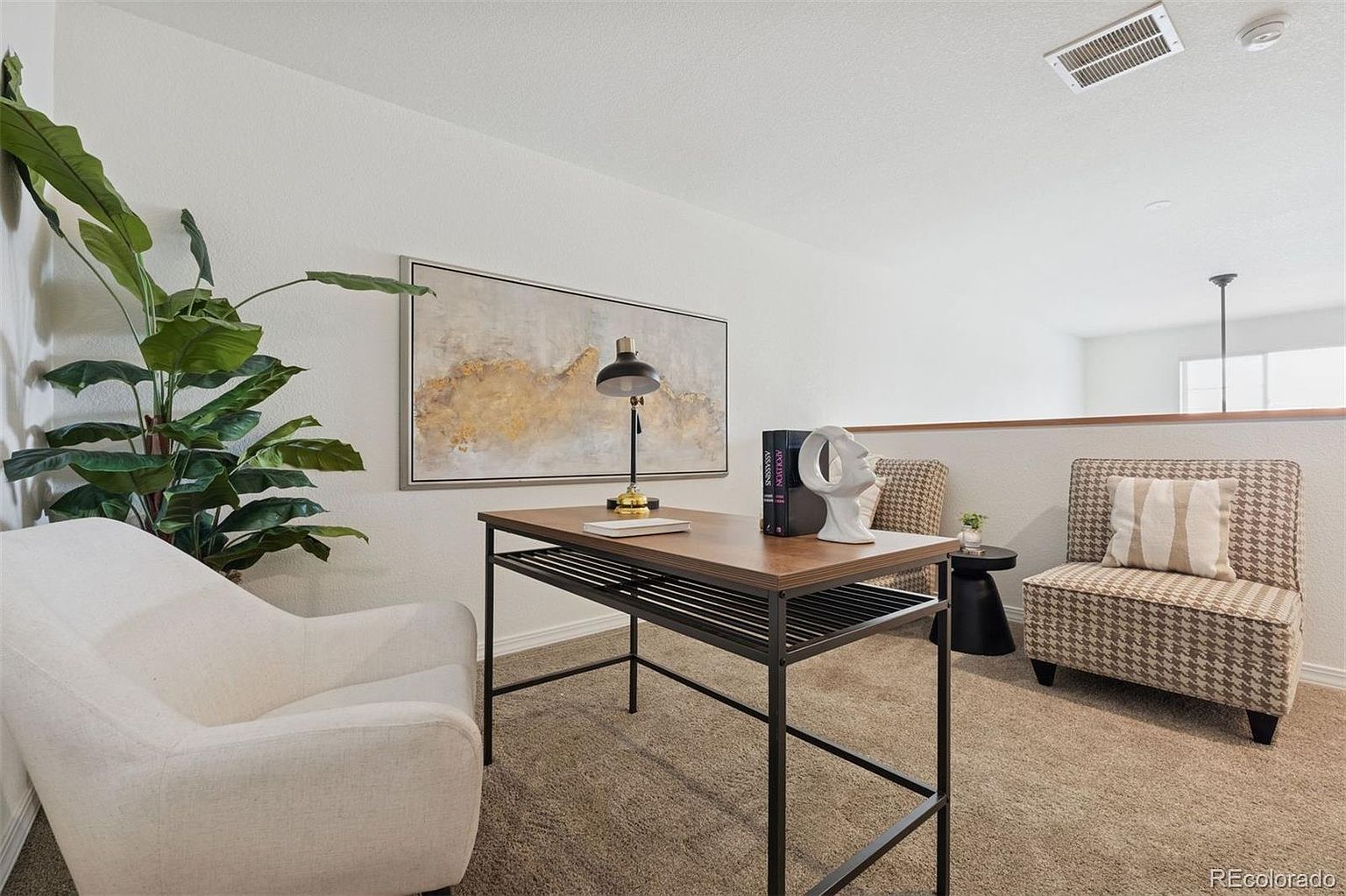 This interior shot showcases a stylish office/study area featuring a wooden desk with a black metal frame, complemented by a modern white chair and a patterned armchair. An abstract painting hangs above the desk, adding an artistic touch to the space. The room is well-lit and appears to be designed for comfort and productivity.