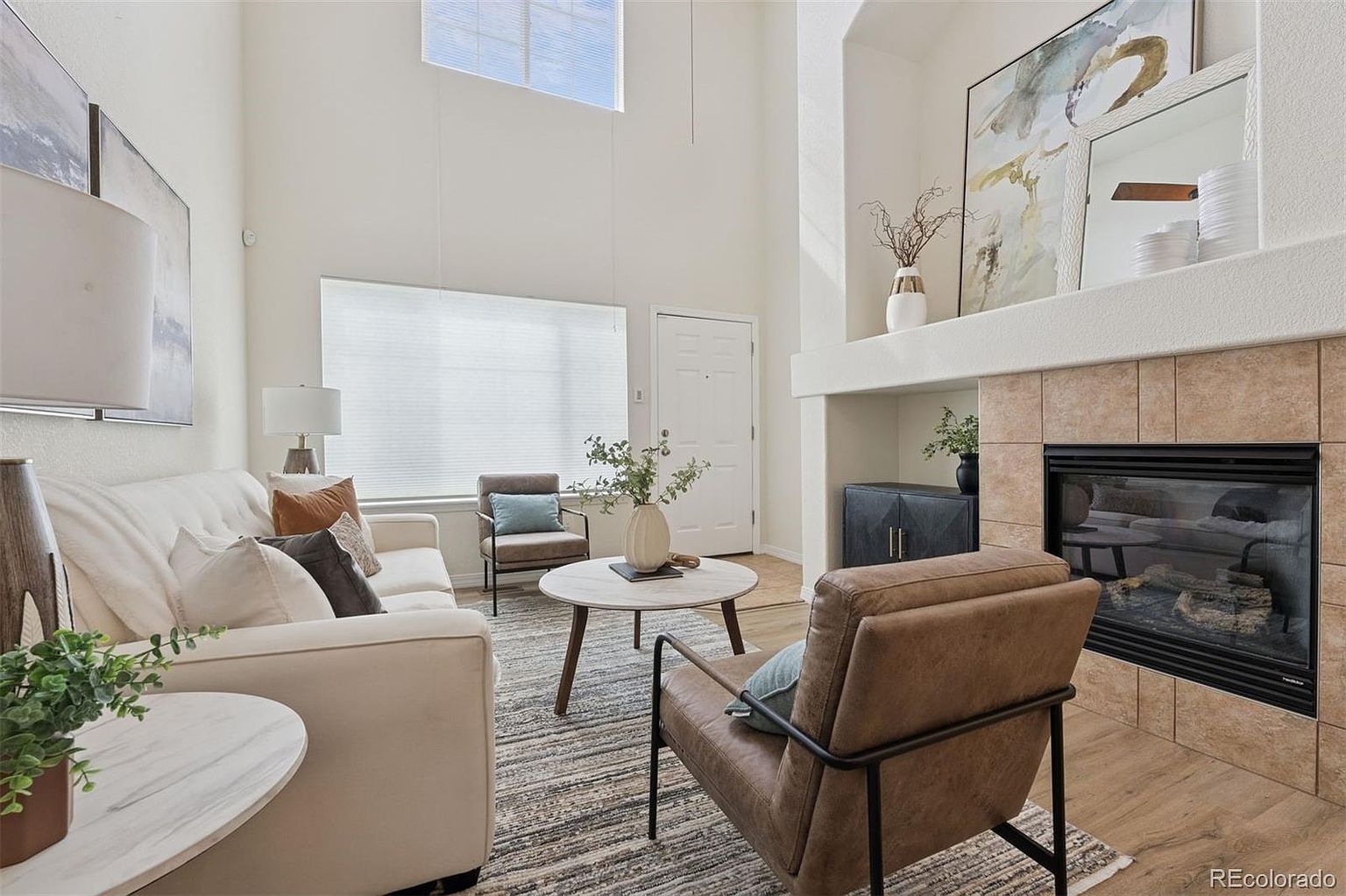 This is an interior shot of a living room featuring a neutral color palette with a fireplace as a focal point. The room is furnished with a light-colored sofa, two accent chairs, and a round coffee table, all arranged on a patterned rug. Natural light streams in through a window, creating a bright and inviting atmosphere.