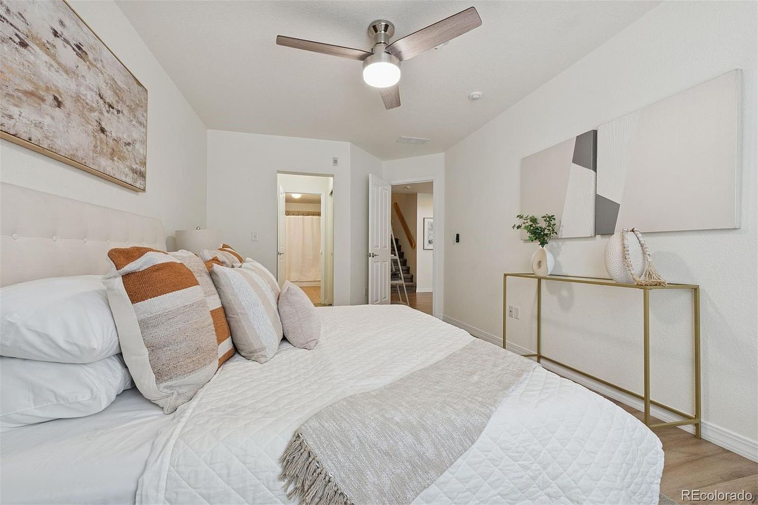 This is a bright and airy primary bedroom featuring a large bed with decorative pillows and a quilted blanket. The room is decorated with modern art and a stylish console table, creating a serene and inviting atmosphere. Natural light fills the space, highlighting the clean lines and neutral color palette.