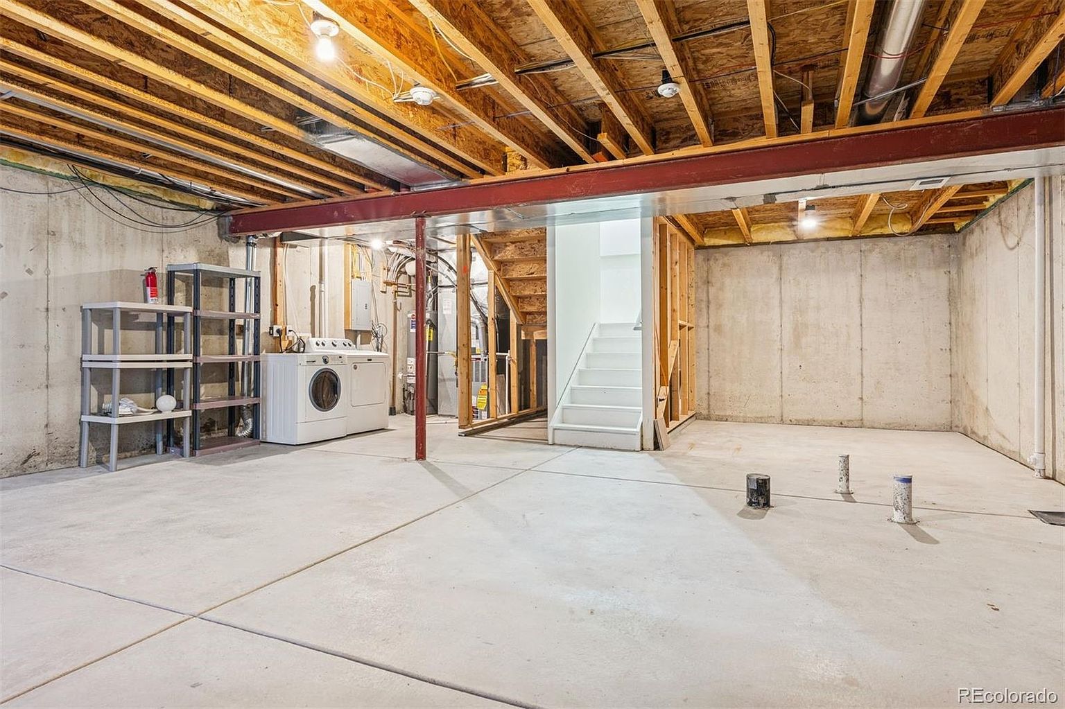 This is a basement interior featuring exposed wooden ceiling beams and a concrete floor. A laundry area with a washer and dryer is visible, along with metal shelving. The space includes unfinished areas with exposed framing and a staircase leading to the upper level, suggesting potential for customization and expansion.