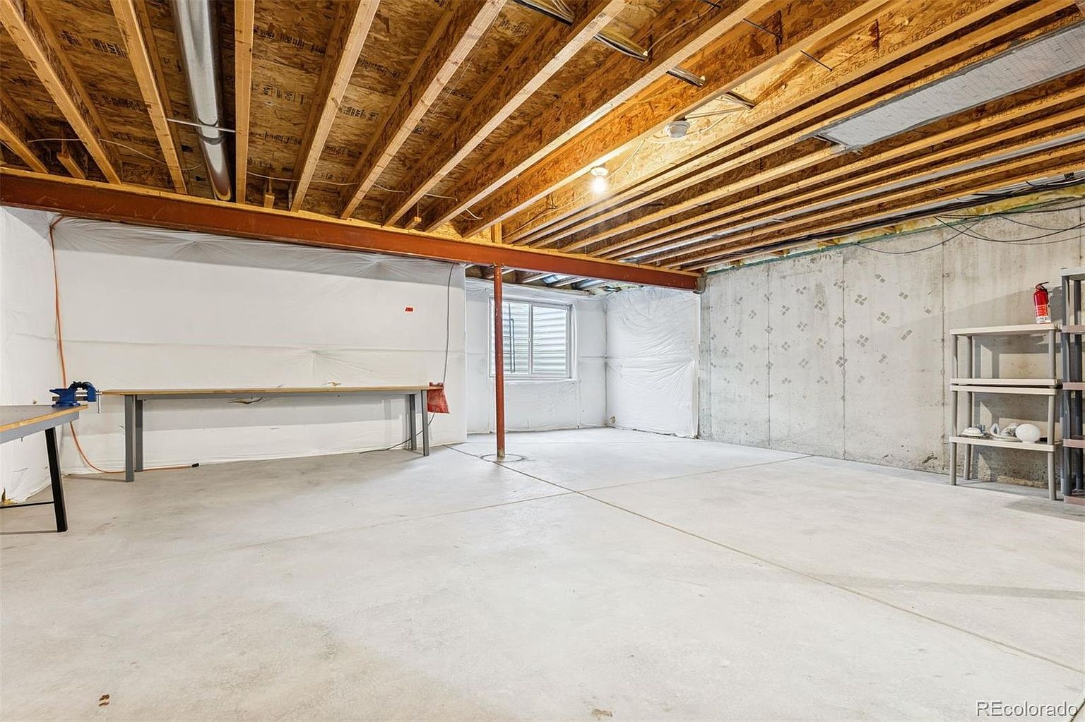 This is an unfinished basement featuring exposed wooden ceiling joists and a concrete floor. A steel support beam runs across the room, supported by a central column. The walls are a mix of concrete and white sheeting, with a window providing natural light. The space appears large and open, offering potential for customization.