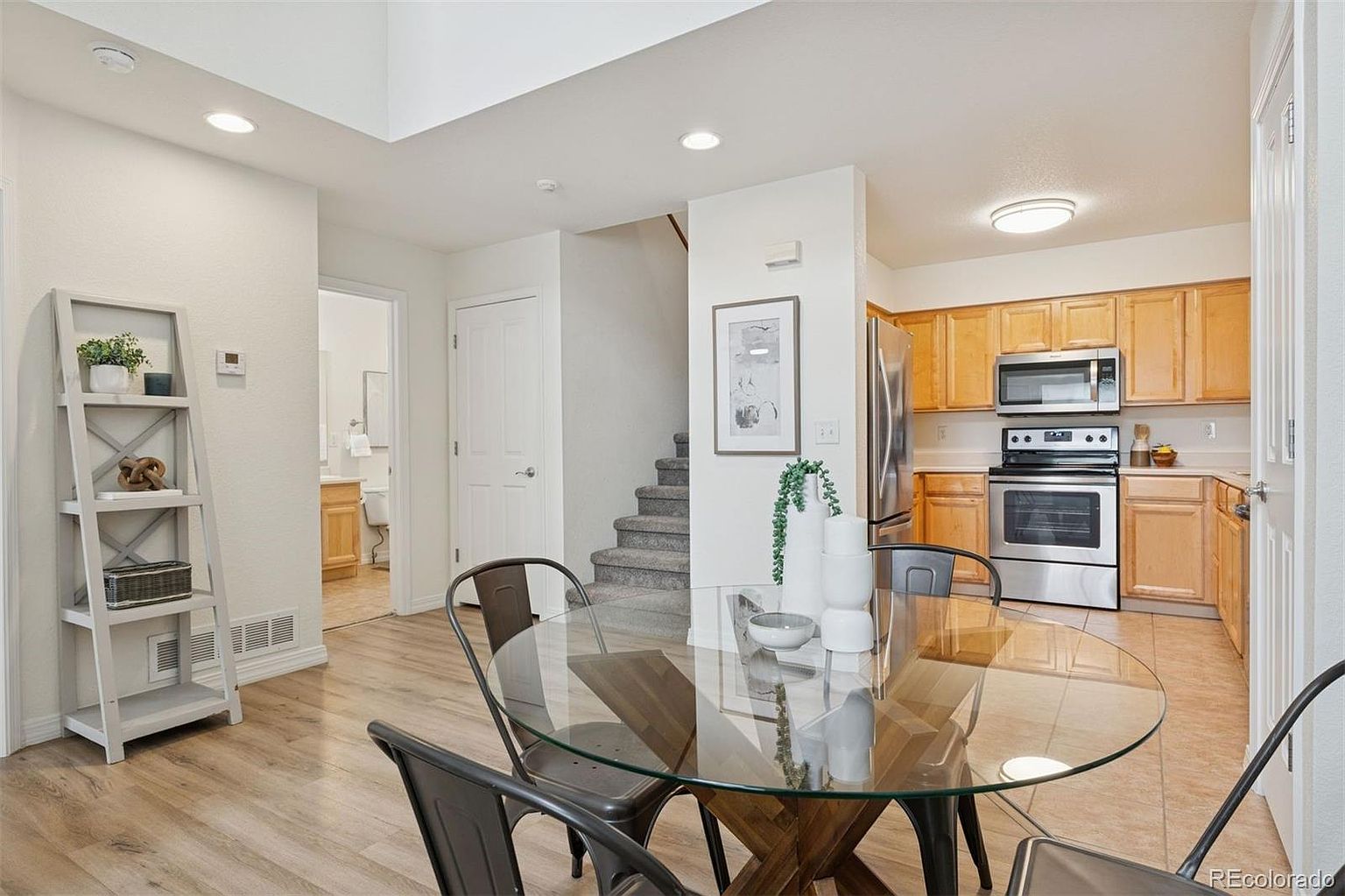 This interior shot showcases a dining area adjacent to a kitchen and stairs. The dining area features a round glass table with metal chairs, set on a light wood floor. The kitchen has wooden cabinets and stainless steel appliances, while the stairs lead to an upper level, creating an open and inviting space.