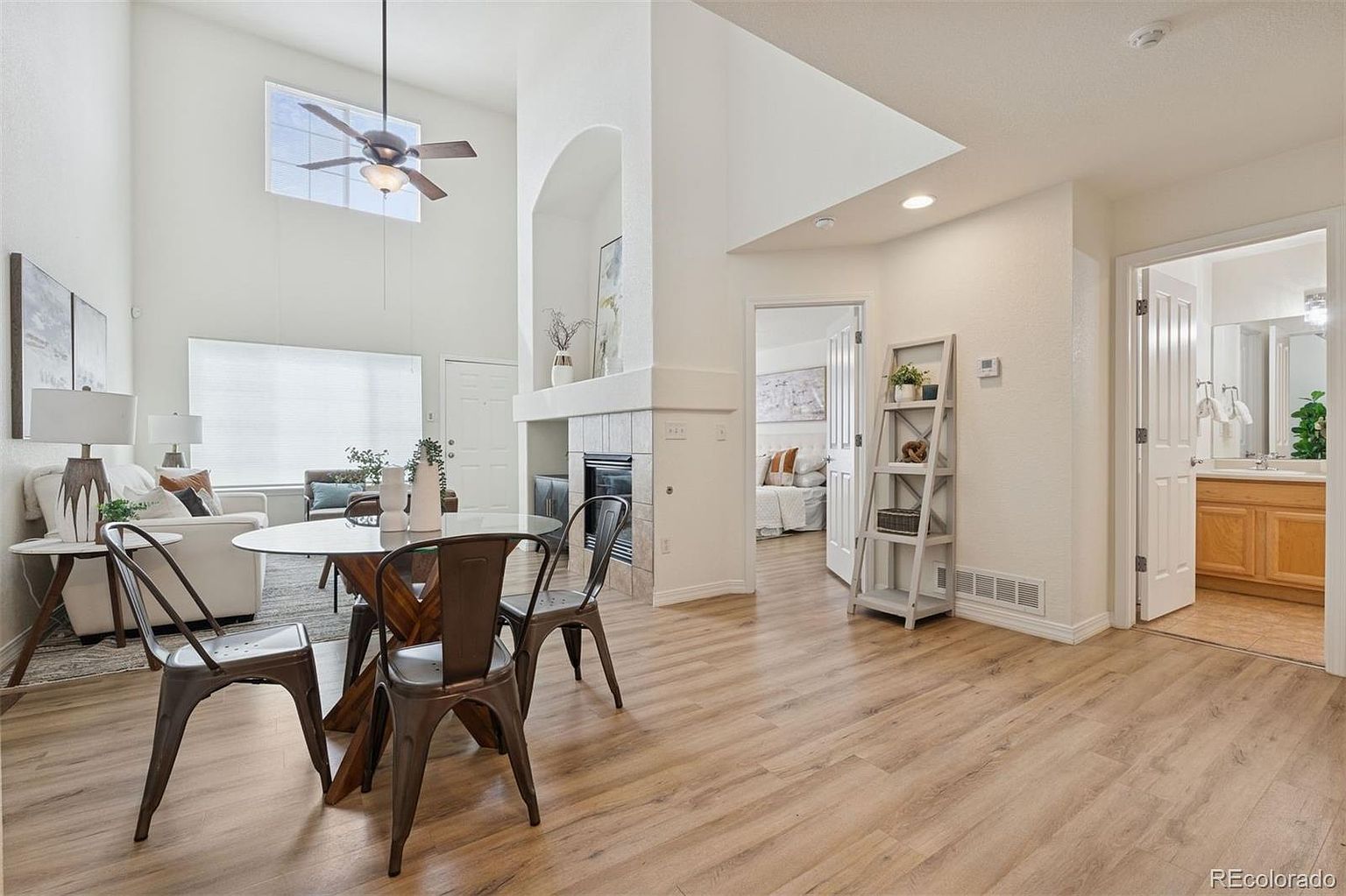 This interior shot showcases a dining area with a round glass-top table and metal chairs, set against a backdrop of light wood flooring and neutral-toned walls. The space is well-lit, with natural light filtering through a window, and features an open layout that connects to other living areas. A fireplace and decorative shelving add to the room's charm and appeal.