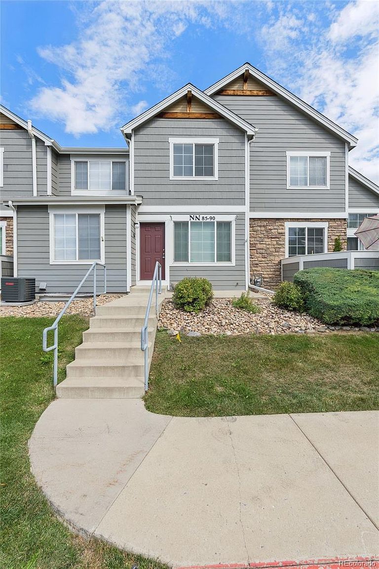 This is a front view of a two-story townhouse with gray siding and a brick accent. A concrete walkway leads to a set of stairs with metal railings, ascending to the front door. The landscaping includes grass, bushes, and decorative rocks, creating a well-maintained and inviting curb appeal.