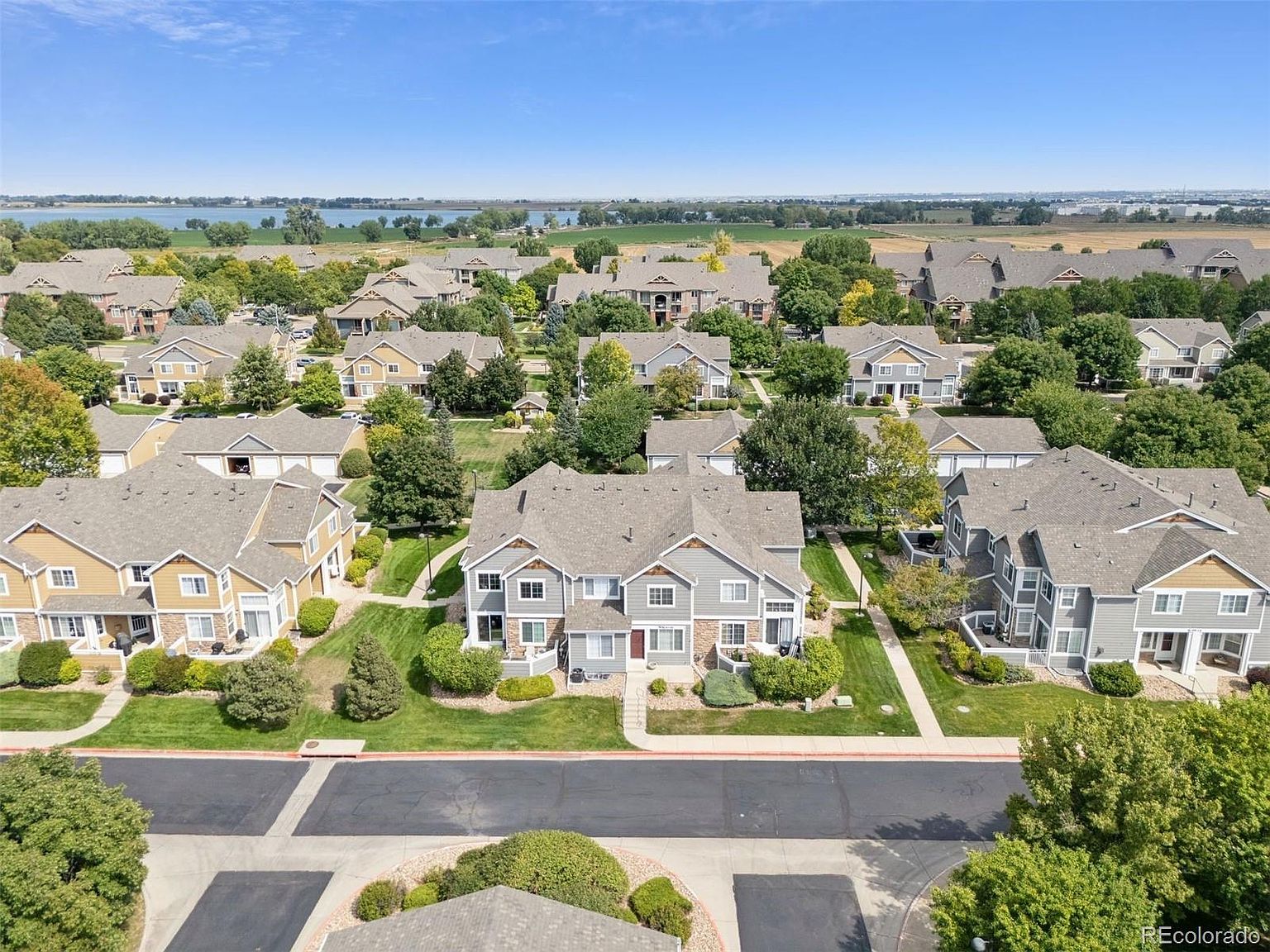 This aerial view showcases a well-maintained residential community with multiple townhouses and condos. The properties feature manicured lawns, mature trees, and paved streets. In the background, a lake and open fields add to the scenic setting, creating a peaceful and desirable neighborhood.