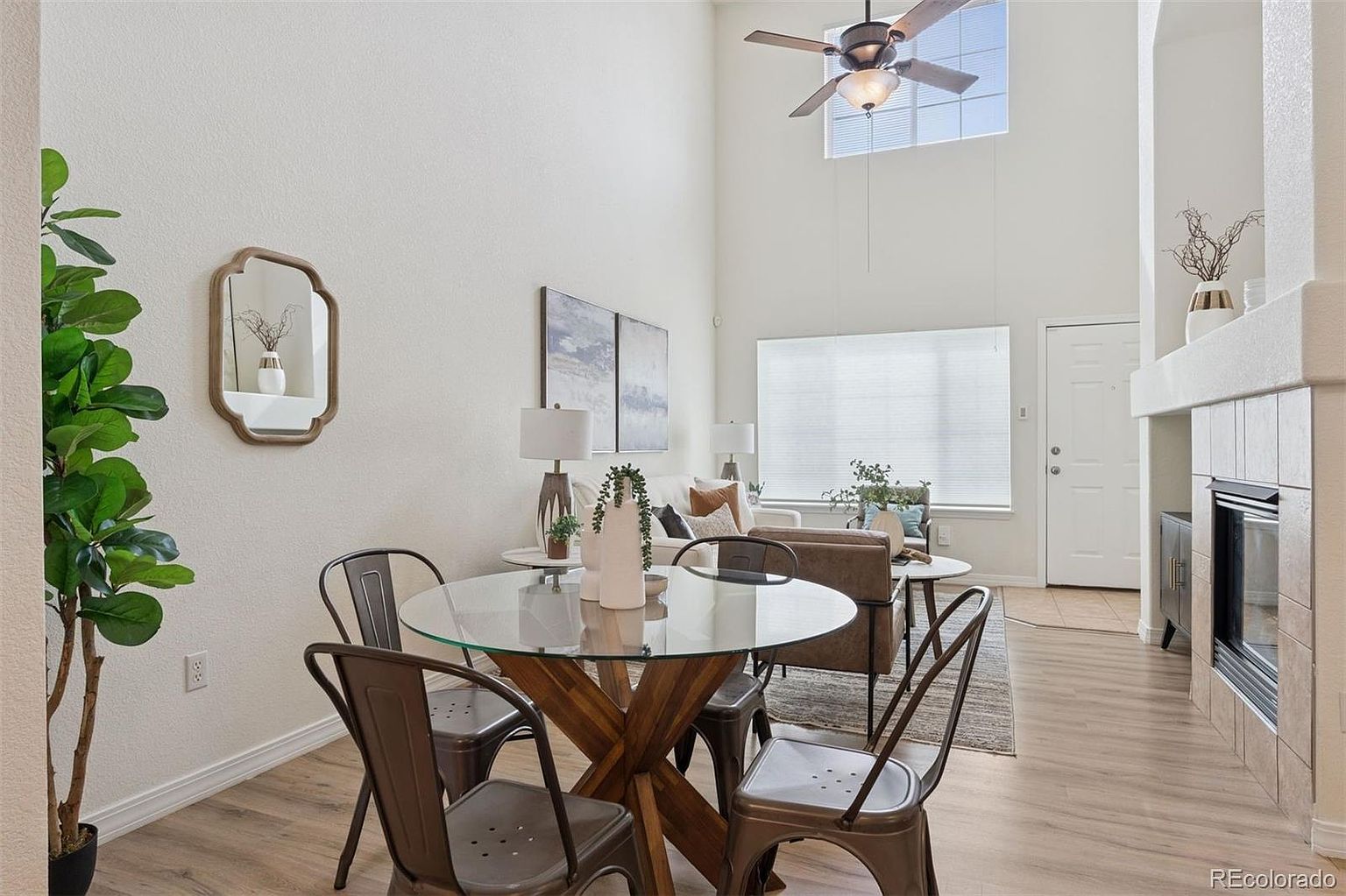 This interior shot showcases a bright and airy living room with a modern aesthetic. The room features a dining area with a glass-top table and metal chairs, a comfortable seating area with a sofa and accent chairs, and a fireplace with tile surround. Natural light floods the space through a large window, highlighting the light wood flooring and neutral color palette.