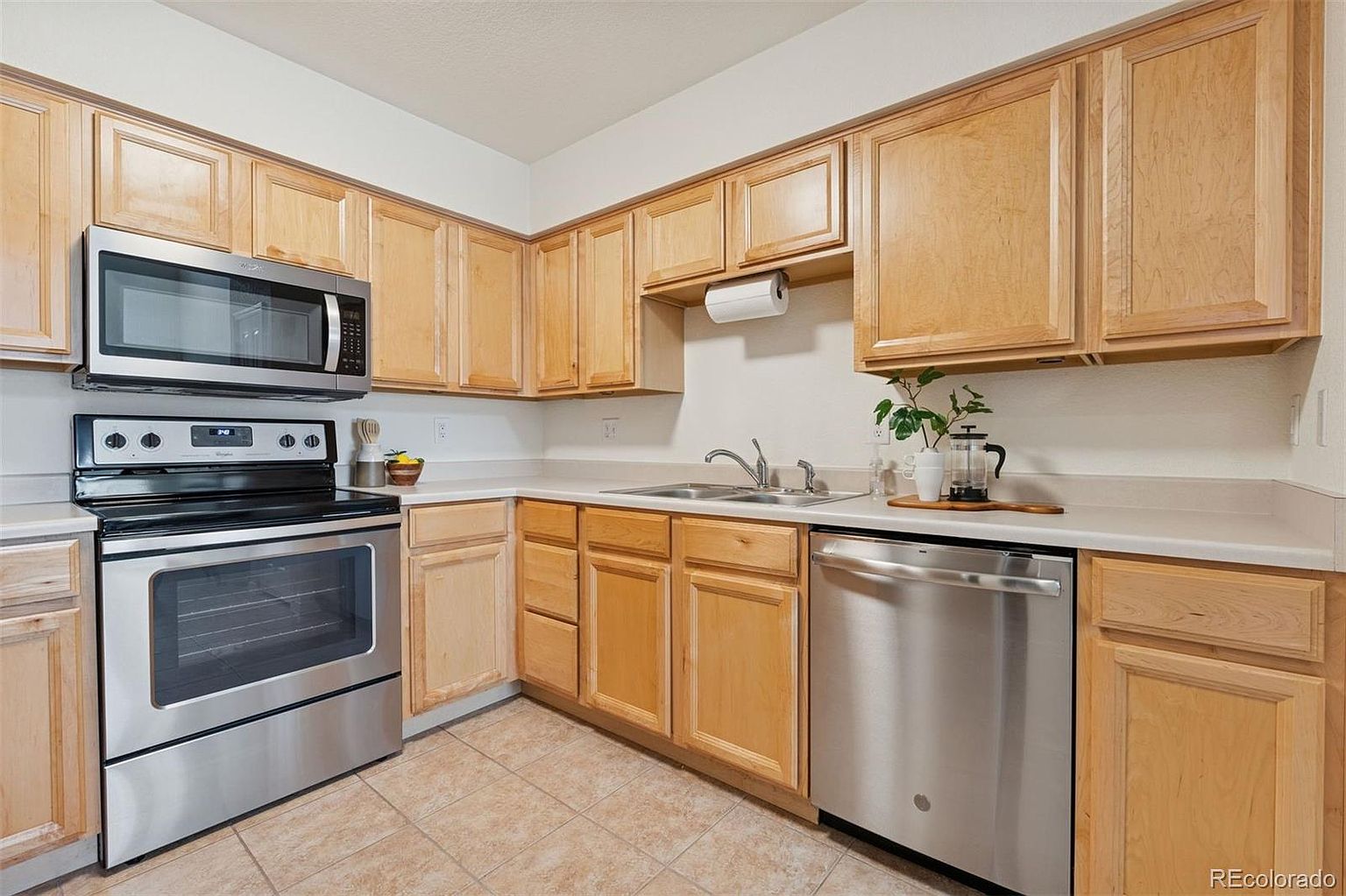 This is a well-lit kitchen featuring light wood cabinetry, stainless steel appliances, and light countertops. The kitchen includes a microwave, oven, dishwasher, and a double basin sink. The flooring is a neutral tile, and the overall impression is clean and functional.