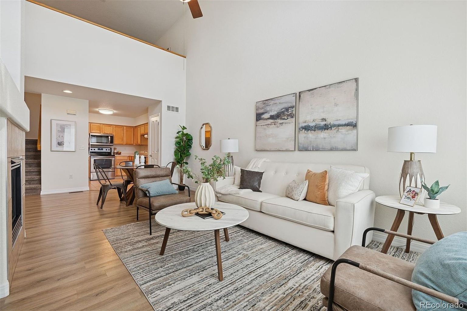 This is an interior shot of a living room featuring a white sofa with decorative pillows, a round coffee table, and two armchairs. The room is decorated with modern artwork and plants, creating a cozy and inviting atmosphere. The flooring is light wood, and a patterned rug anchors the seating area.