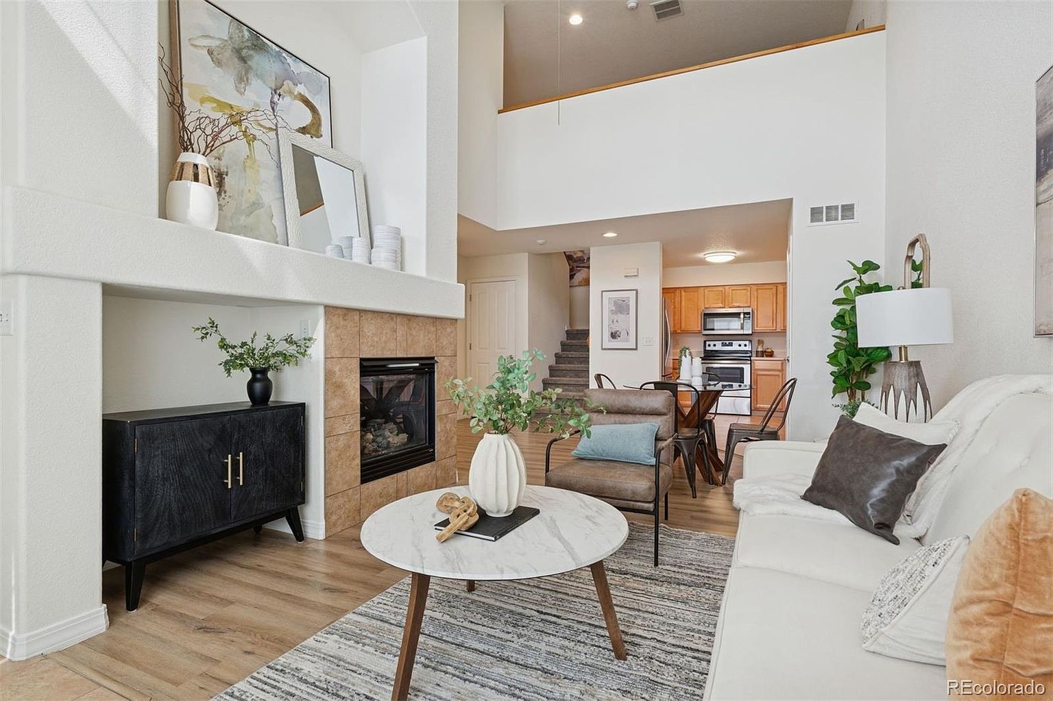 This is an interior shot of a living room featuring a fireplace with a decorative mantel, a dark wood cabinet, and a round marble-topped coffee table. A white sofa and a leather armchair provide seating, and a patterned rug anchors the space. The room has a high ceiling and an open layout, leading into the kitchen and dining area.