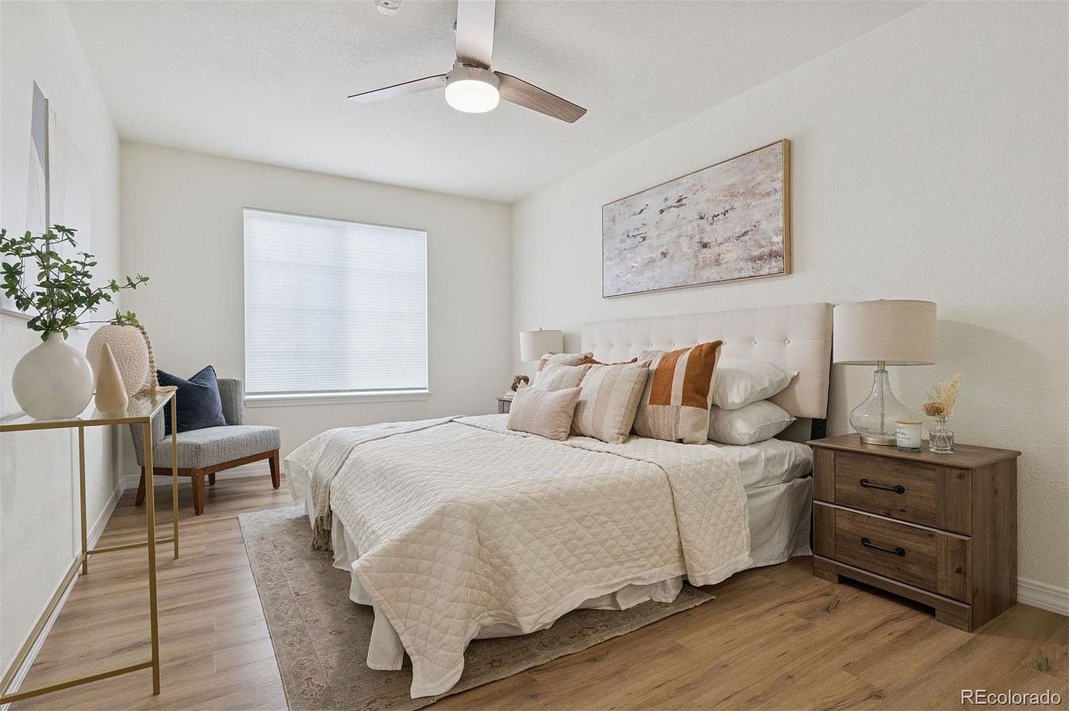 This is a well-lit primary bedroom featuring a neutral color palette. The room includes a bed with a tufted headboard, several decorative pillows, and a quilted blanket. A wooden nightstand with a lamp sits beside the bed, and a piece of artwork hangs above the headboard, contributing to the room's serene and inviting atmosphere.
