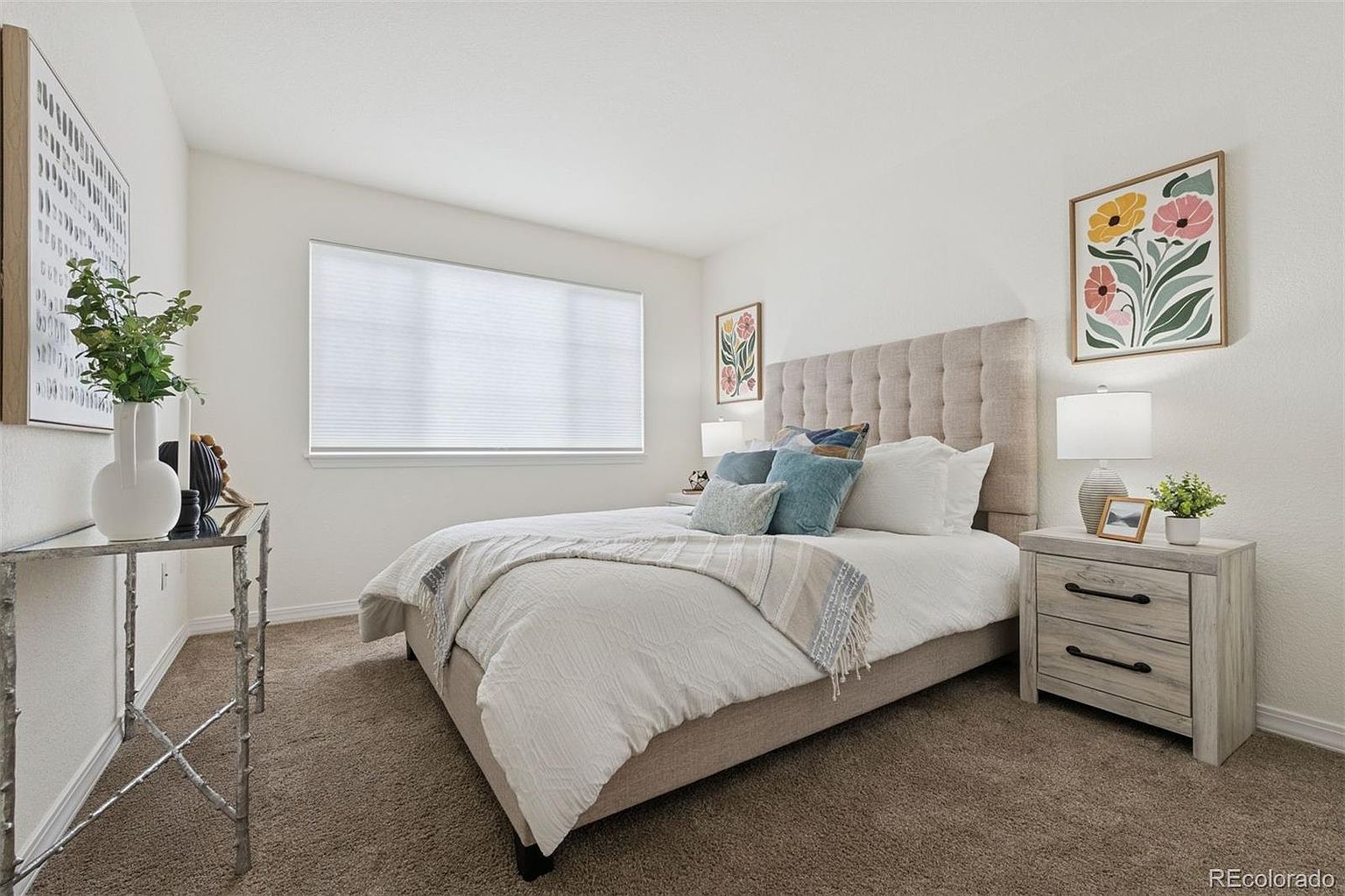This is a well-staged primary bedroom featuring a neutral color palette. The bed has a tufted headboard and is adorned with decorative pillows and a throw blanket. A nightstand with a lamp and decorative items sits beside the bed, and artwork hangs on the wall, creating a serene and inviting atmosphere.