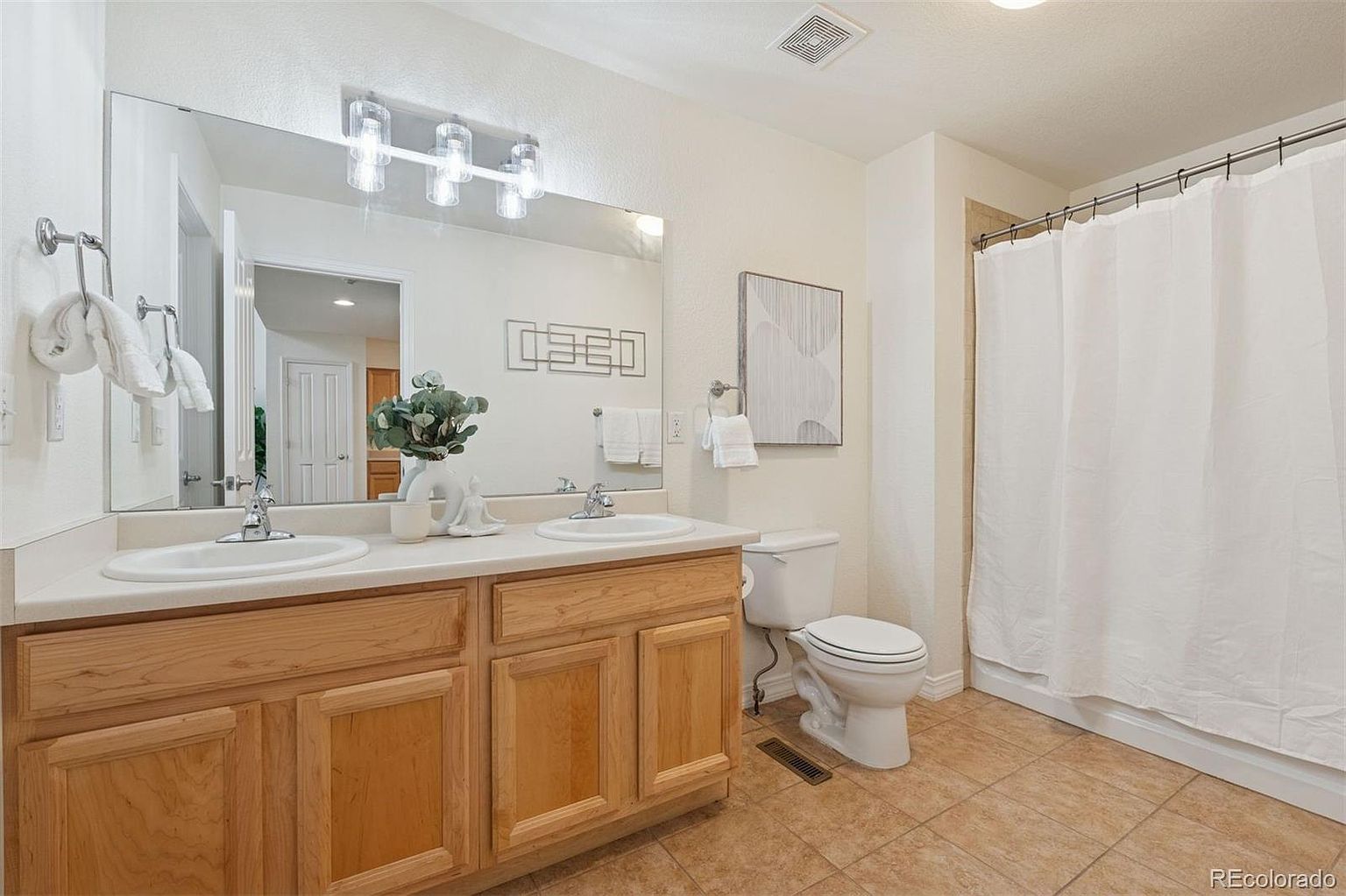 This is a well-lit bathroom featuring a double vanity with light wood cabinets and a white countertop. A large mirror hangs above the sinks, reflecting the interior of the home. The bathroom also includes a toilet and a shower with a white curtain, all set against a backdrop of neutral-colored walls and tiled flooring.