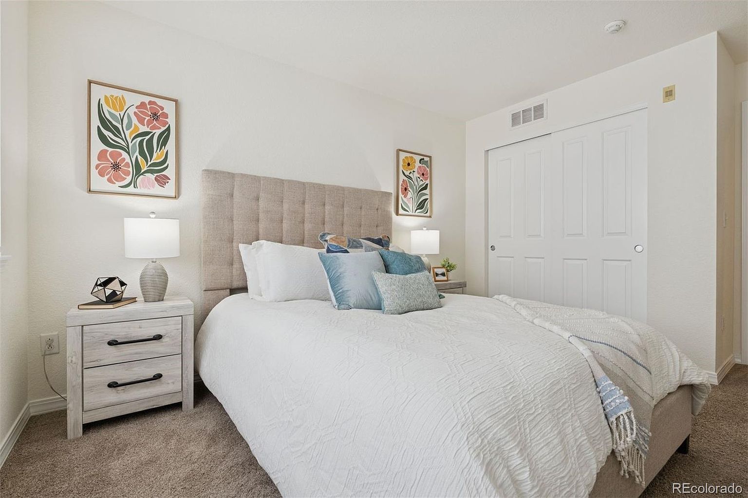 This is a well-staged primary bedroom featuring a neutral color palette. The bed has a tufted headboard and is adorned with white bedding and decorative pillows. A nightstand with a lamp and decorative items sits beside the bed, and artwork hangs on the wall, creating a cozy and inviting atmosphere.
