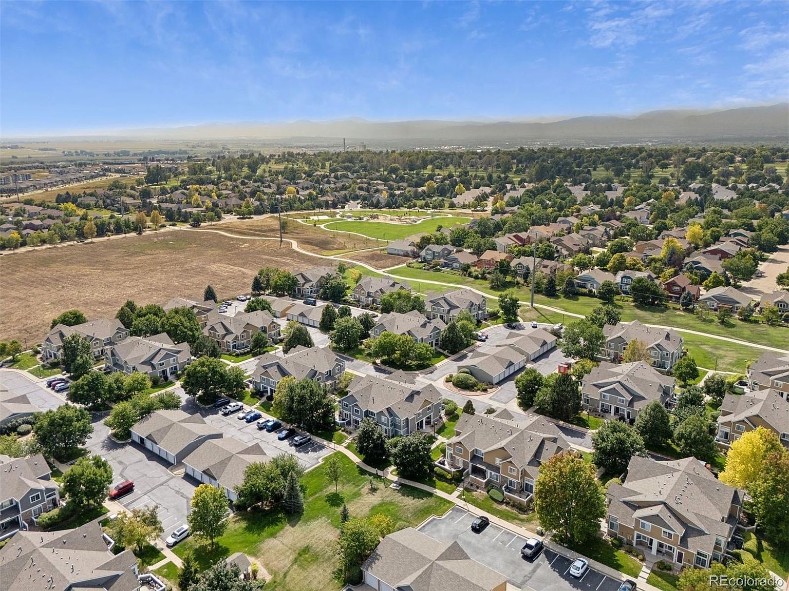 This aerial view showcases a well-maintained residential neighborhood with a mix of townhouses and single-family homes. Lush green spaces and mature trees are interspersed throughout the community, with a park and open field visible in the background. The overall impression is one of a peaceful and established suburban setting.