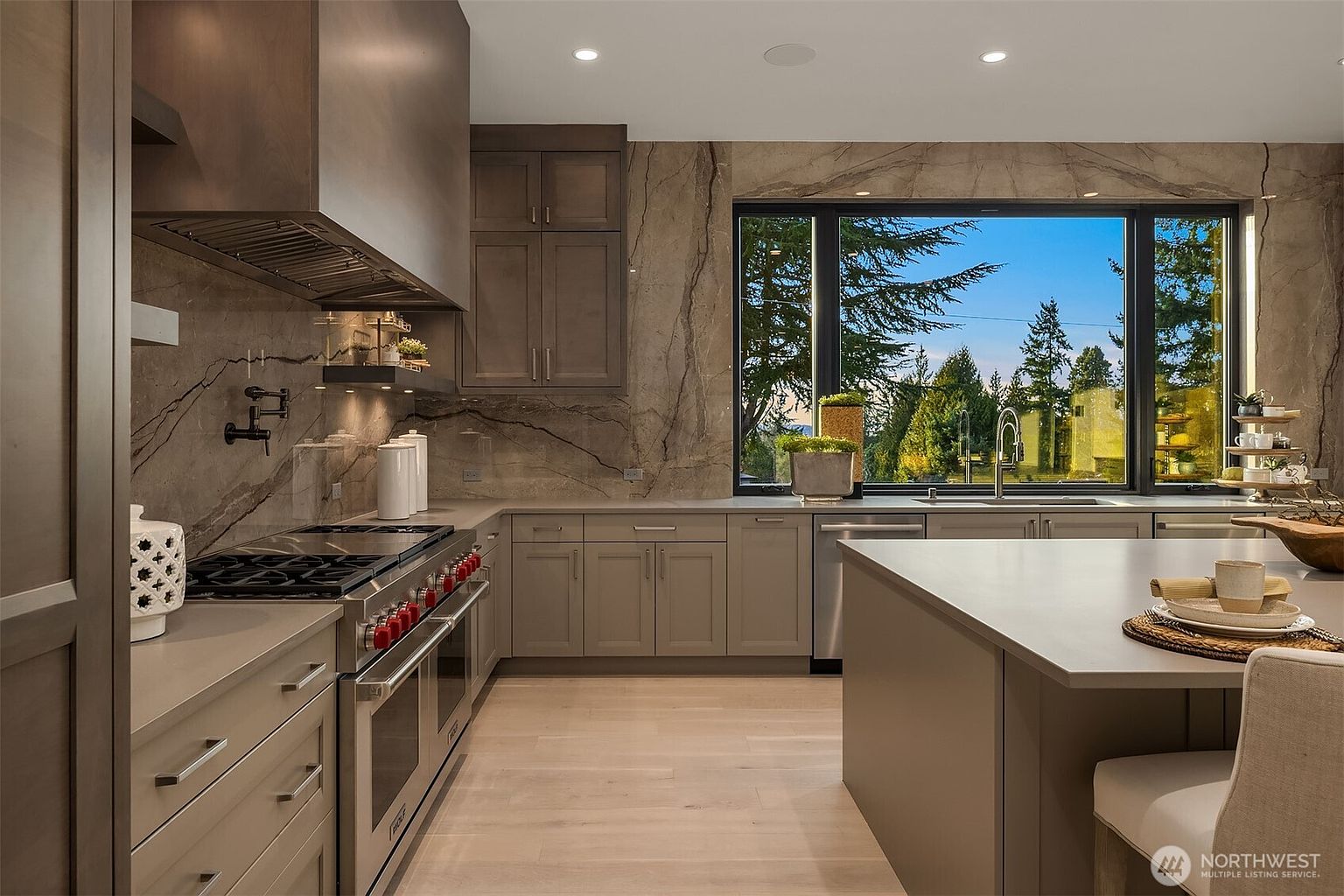 This modern kitchen features sleek, neutral-toned cabinetry paired with a large central island and light wood flooring. A striking stone backsplash extends behind the professional-grade range and hood, while a large window above the sink provides a clear view of the outdoor landscape. The space is illuminated by recessed lighting, creating a sophisticated and functional atmosphere.