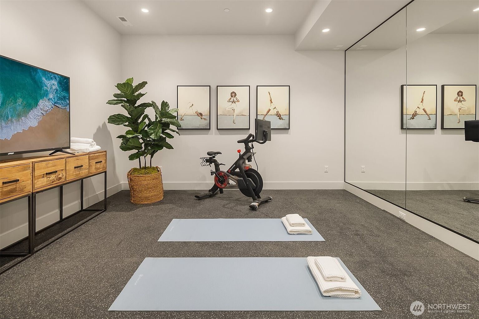 This well-appointed home gym features a Peloton exercise bike centered in the room, flanked by a large potted plant and a rustic wooden console table topped with a television. Two light blue yoga mats with folded white towels are laid out on the dark carpet, while a large wall mirror on the right enhances the sense of space. The walls are adorned with three framed yoga-themed art prints, creating a serene and focused atmosphere for fitness.