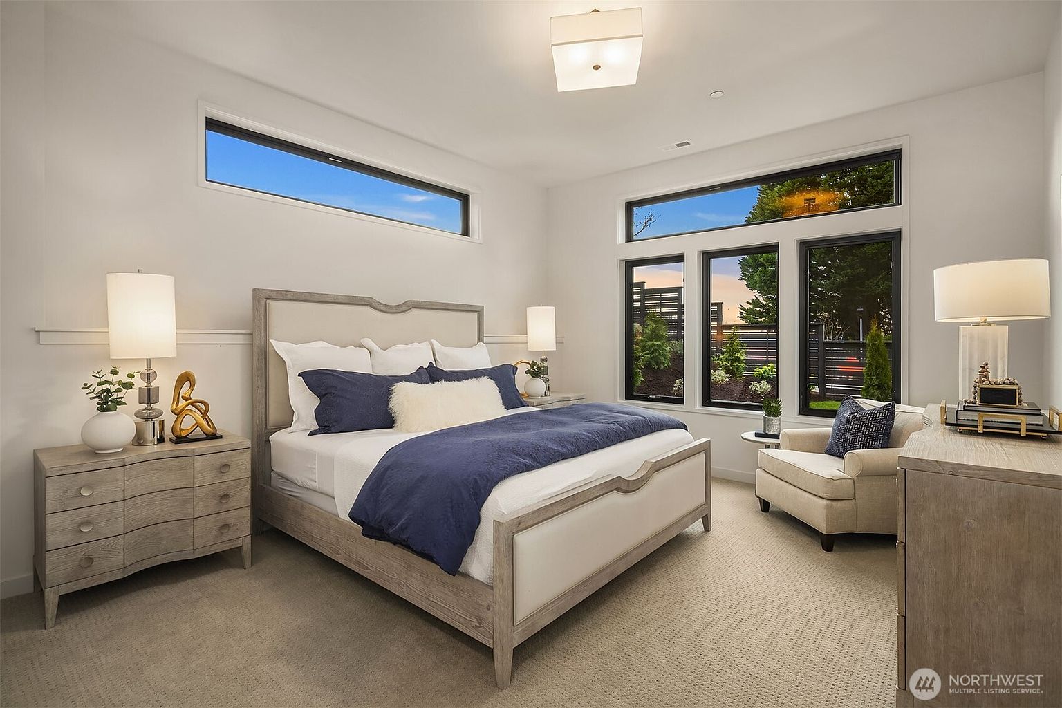 This primary bedroom features a neutral color palette with a light wood-framed bed, a navy blue duvet, and matching accent pillows. The room is illuminated by a central ceiling fixture and two bedside lamps, while large windows and a horizontal transom window provide ample natural light and views of the exterior. A comfortable armchair sits in the corner, creating a cozy reading nook, and the space is finished with light-toned wood furniture and plush carpeting.