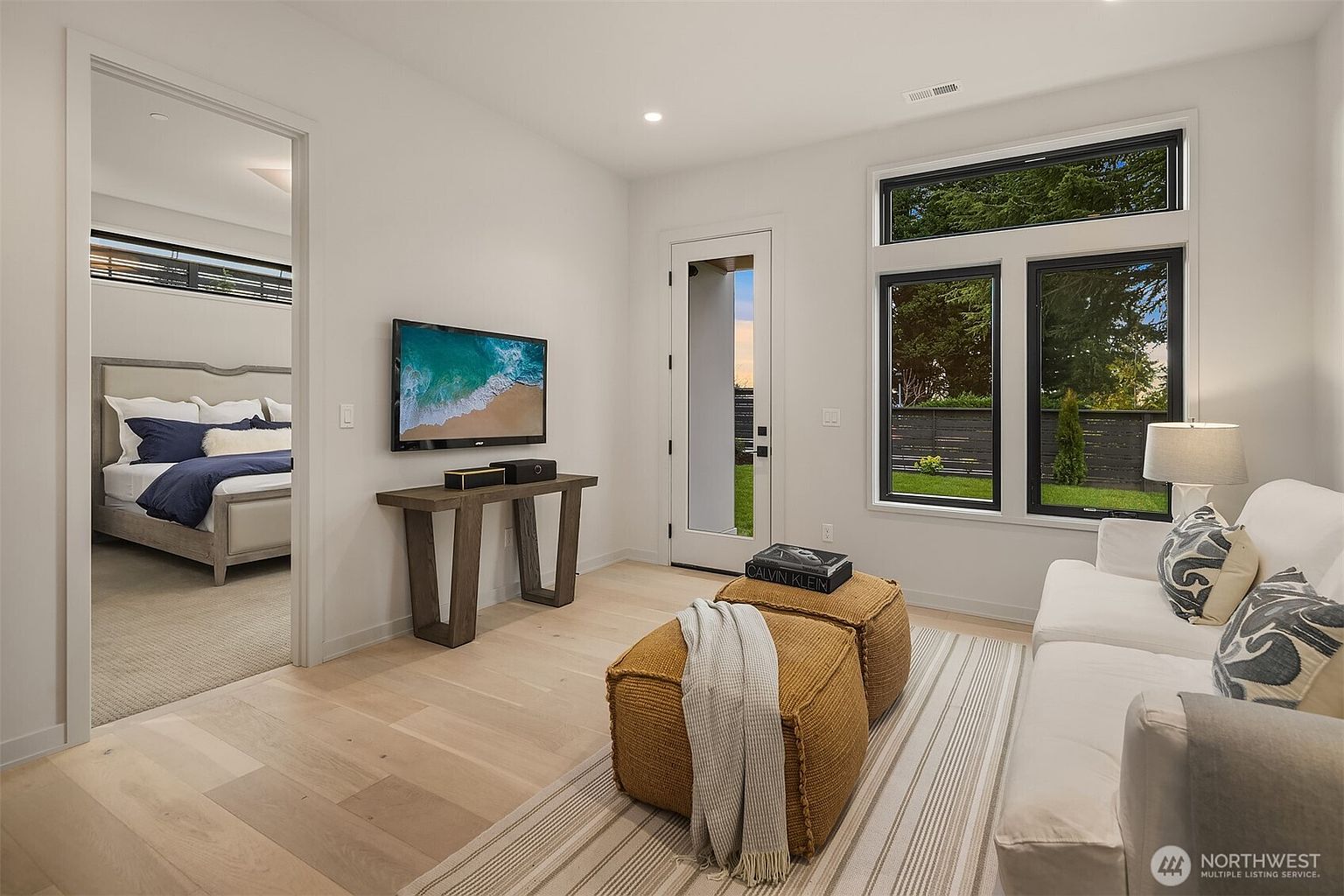 This bright and modern living room features light wood flooring, a white sofa, and two textured mustard-colored ottomans centered on a striped area rug. A wall-mounted television sits above a console table, while a glass-paned door and large windows provide natural light and a view of the backyard. The room offers a seamless transition into an adjacent bedroom, creating an open and airy atmosphere.
