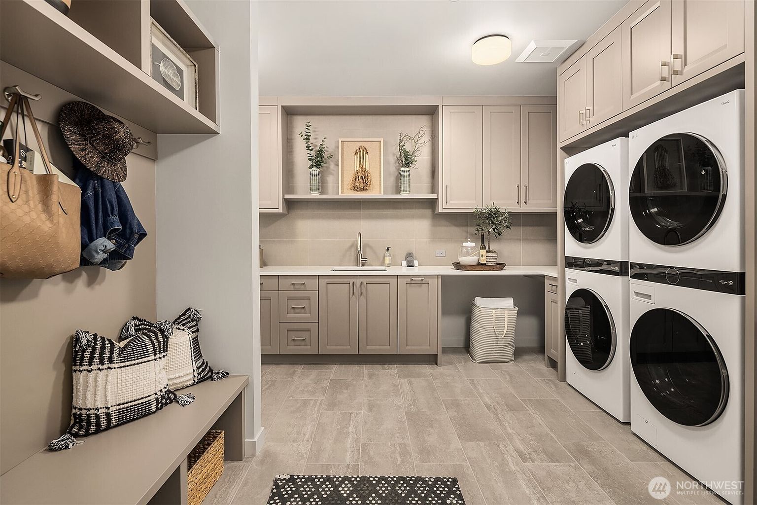 This well-appointed laundry room features a functional mudroom area with a built-in bench, coat hooks, and overhead storage. The space includes a stacked washer and dryer set, a sink with a countertop workspace, and neutral-toned cabinetry that provides ample storage. The room is finished with light-colored tile flooring and decorative accents, creating a clean, organized, and inviting atmosphere.