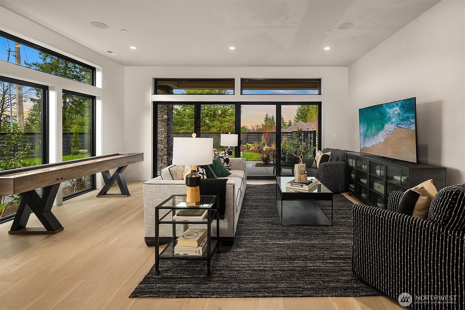 This contemporary living room features light wood flooring, a large dark area rug, and a neutral-toned sofa paired with patterned armchairs. A shuffleboard table sits to the left, while a large television is mounted above a dark media console on the right. The space is brightened by expansive glass doors that provide a clear view of the backyard, creating a seamless transition between indoor and outdoor living areas.