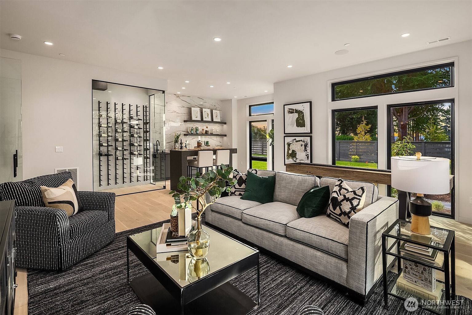 This sophisticated living room features a contemporary design with a gray sofa, a striped armchair, and a central coffee table set atop a dark textured rug. To the left, a glass-enclosed wine display adds a luxurious touch, while a bar area with floating shelves and stone-patterned walls sits in the background. Large windows provide ample natural light and a view of the outdoor greenery, creating an open and inviting atmosphere.