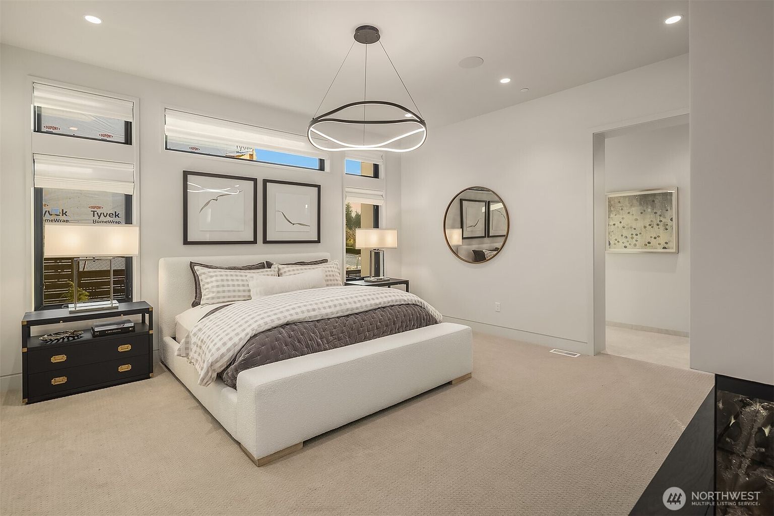 This spacious primary bedroom features a contemporary aesthetic with a large, upholstered bed as the central focal point, flanked by two dark-toned nightstands with matching lamps. The room is illuminated by a striking, modern circular chandelier and benefits from natural light streaming through large windows adorned with white shades. A circular wall mirror adds a decorative touch, while an open doorway leads into an adjacent space, creating an airy and open floor plan.