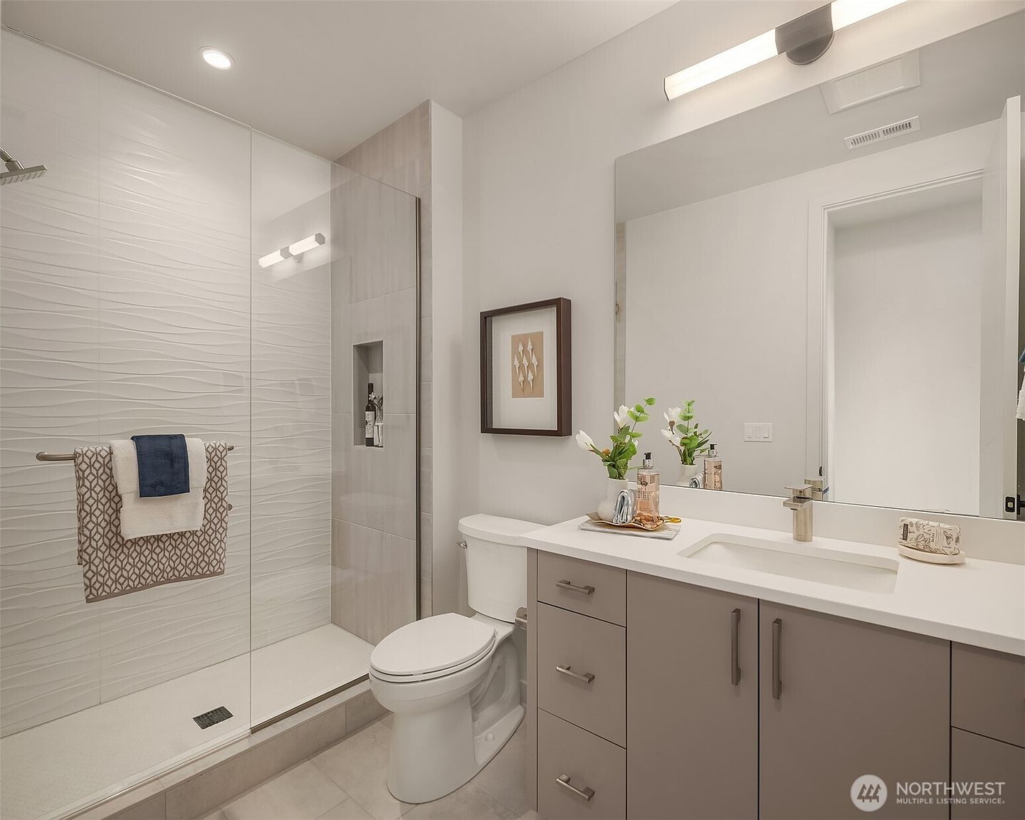 This modern bathroom features a sleek vanity with a light-toned countertop and gray cabinetry, paired with a large, frameless mirror. To the left, a walk-in shower is enclosed by a glass panel and showcases textured, wavy-patterned wall tiles. The space is bright and clean, accented by contemporary fixtures and minimalist decor.