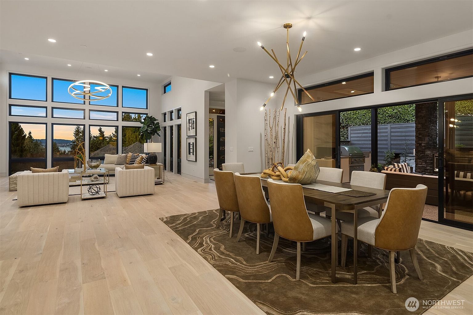 This open-concept living and dining area features light wood flooring and a sophisticated, modern aesthetic. The dining space is anchored by a large table with upholstered chairs and a striking, multi-armed gold chandelier, while the adjacent living area is bathed in natural light from expansive windows that offer a scenic view. The space flows seamlessly toward an outdoor patio, creating an airy and inviting atmosphere perfect for entertaining.