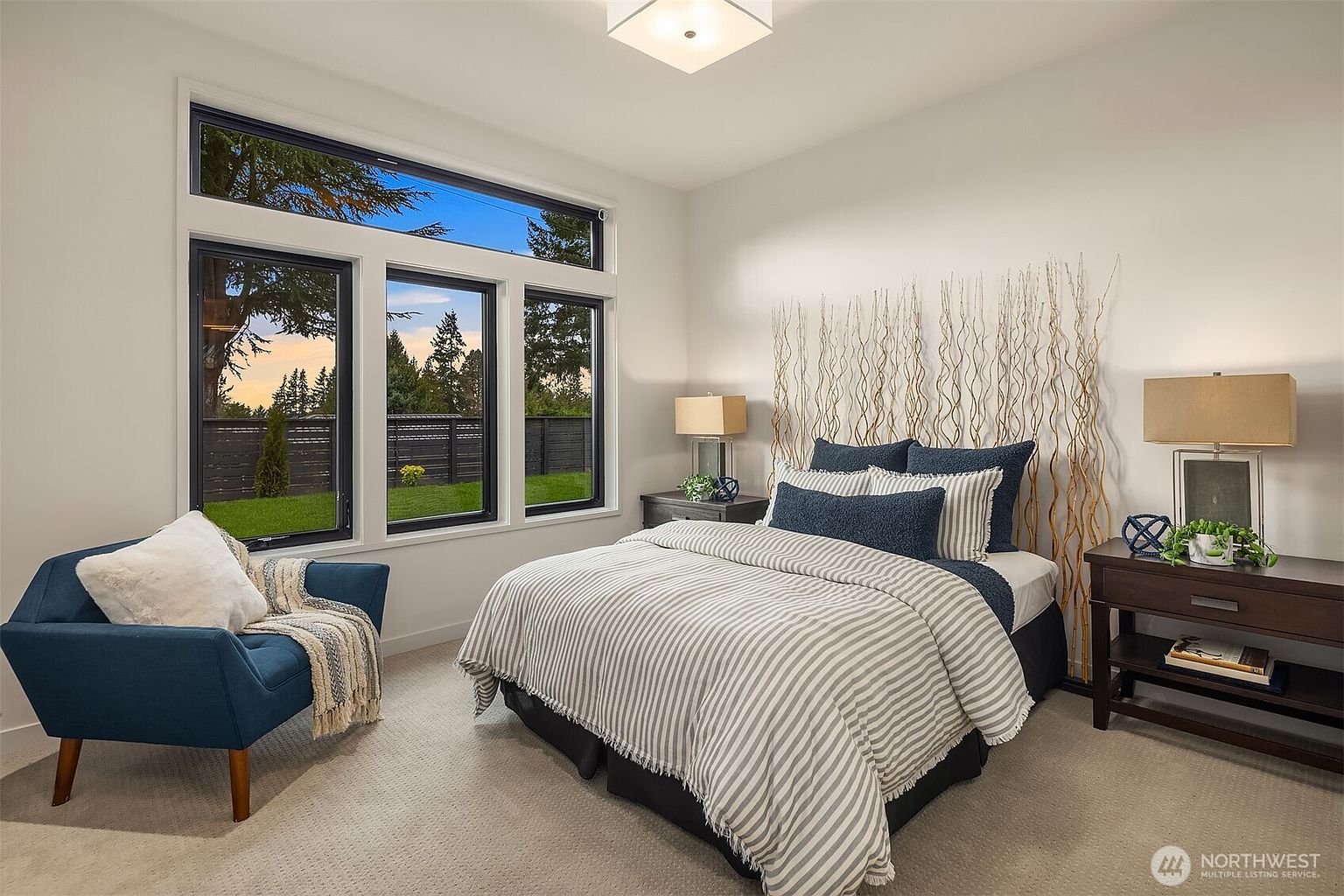 This bright and modern bedroom features a central bed dressed in striped bedding, flanked by two dark wood nightstands with matching lamps. A decorative arrangement of tall, thin branches serves as a unique headboard accent against the neutral wall. To the left, a blue armchair provides a cozy seating area near a large window assembly that offers a view of the backyard.