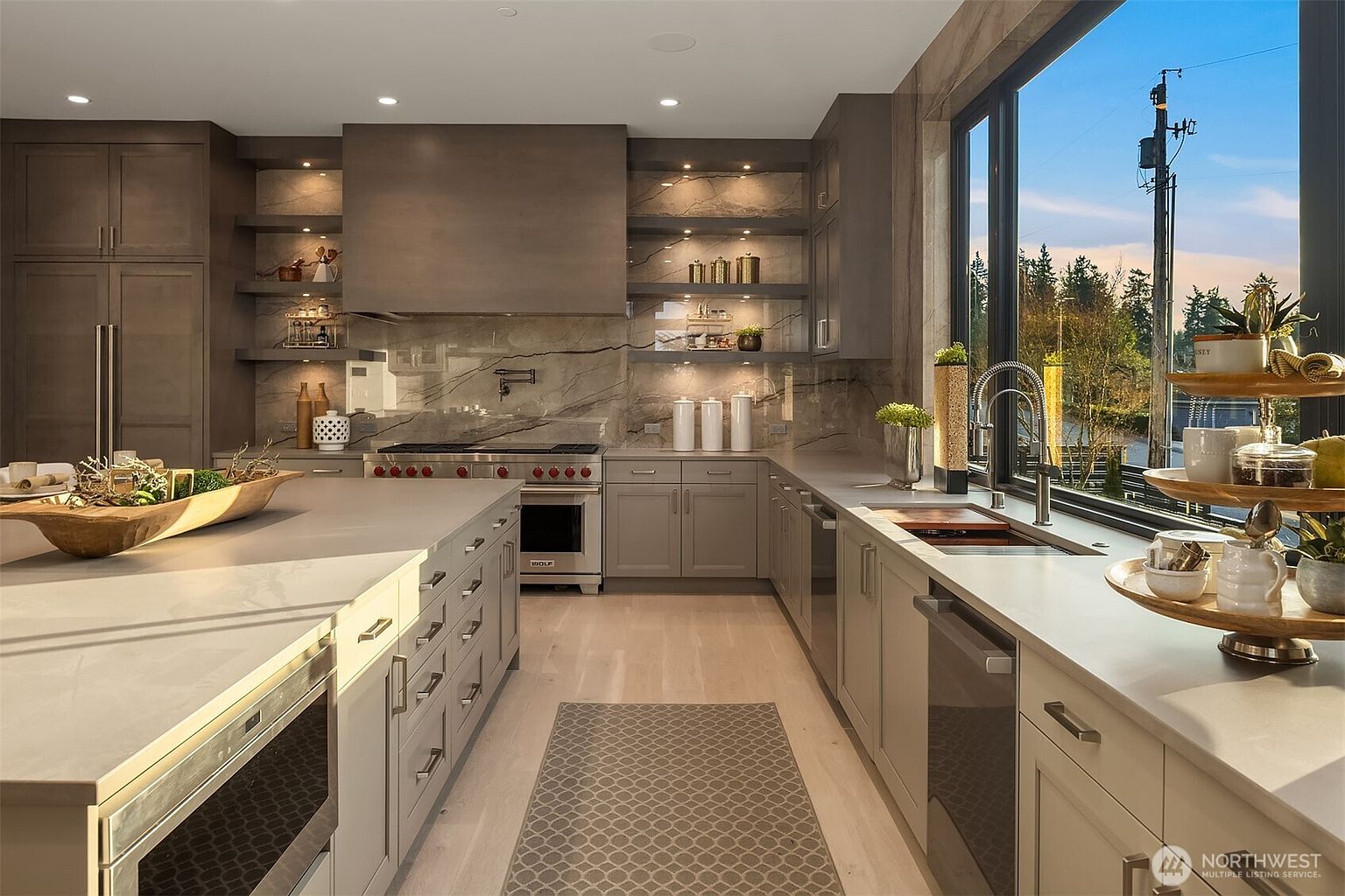 This sophisticated, modern kitchen features sleek, neutral-toned cabinetry and a large central island with a wooden decorative bowl. The space is anchored by a professional-grade range with a custom hood and a stone backsplash that extends to the shelving units. A large window above the sink provides ample natural light and a view of the outdoors, creating an airy and high-end atmosphere.