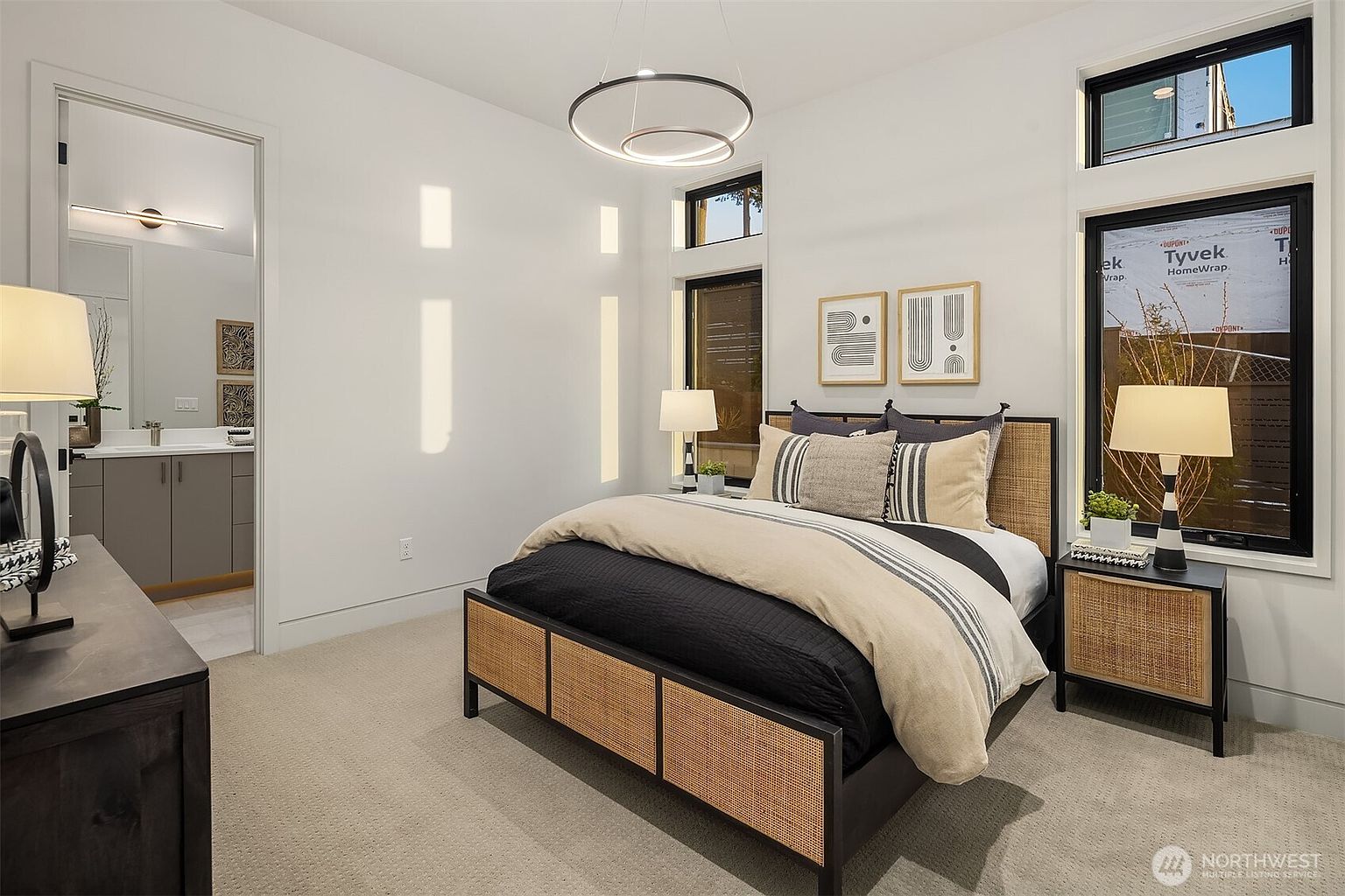 This modern bedroom features a contemporary bed frame with woven cane detailing and a neutral-toned bedding ensemble. The room is illuminated by a unique circular pendant light and two bedside lamps, with large windows providing natural light. An open doorway leads into an adjacent bathroom, creating a cohesive and airy feel throughout the space.