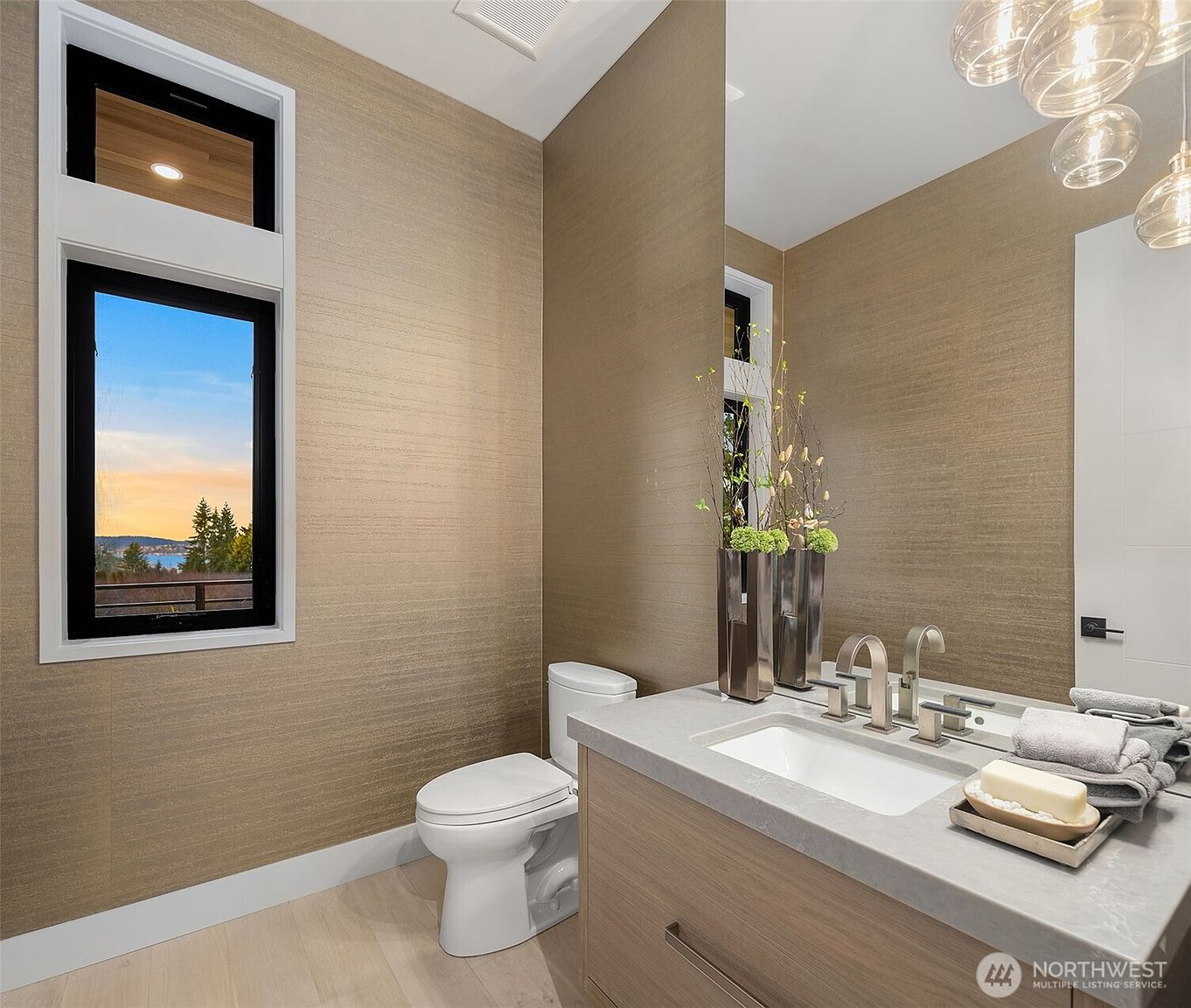 This modern powder room features textured, metallic-toned wallpaper and a floating vanity with a light wood finish and a stone countertop. A large, frameless mirror reflects the space, while a window provides natural light and a scenic view of the outdoors. The room is accented by contemporary brushed-nickel fixtures and a cluster of glass pendant lights.