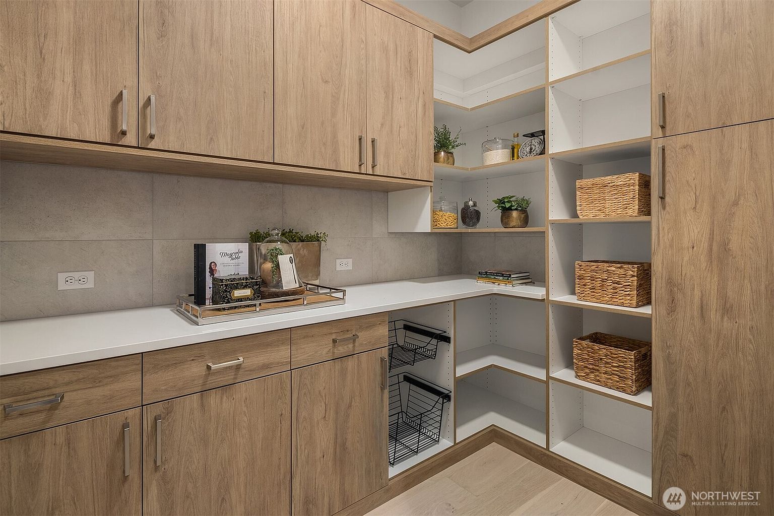 This organized pantry features light wood cabinetry with clean lines and ample storage space, including open shelving and pull-out wire baskets. A white countertop provides a functional workspace, complemented by a neutral tiled backsplash and integrated electrical outlets. The design is modern and efficient, offering a bright and tidy storage solution for a home.
