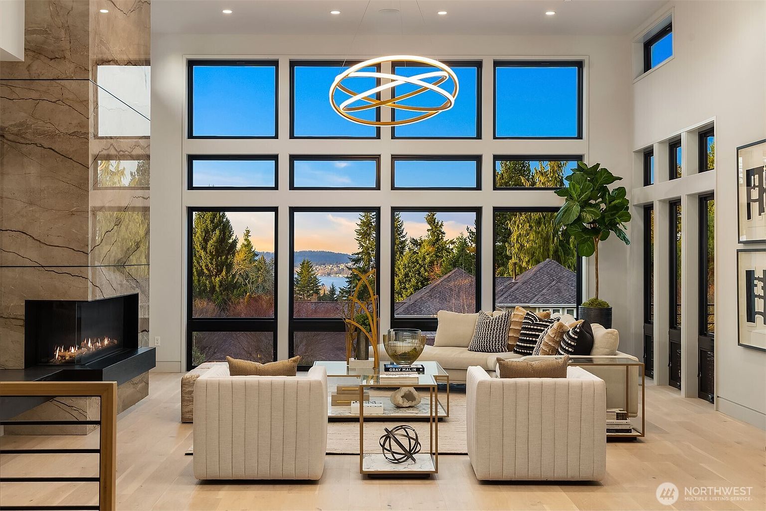 This sophisticated living room features a striking stone-clad chimney breast and a large, circular modern chandelier that serves as a focal point. The space is anchored by a neutral-toned sofa and two matching armchairs, all arranged around a glass-topped coffee table. Expansive windows provide a panoramic view of the surrounding trees and water, filling the room with natural light and creating a seamless connection to the outdoors.