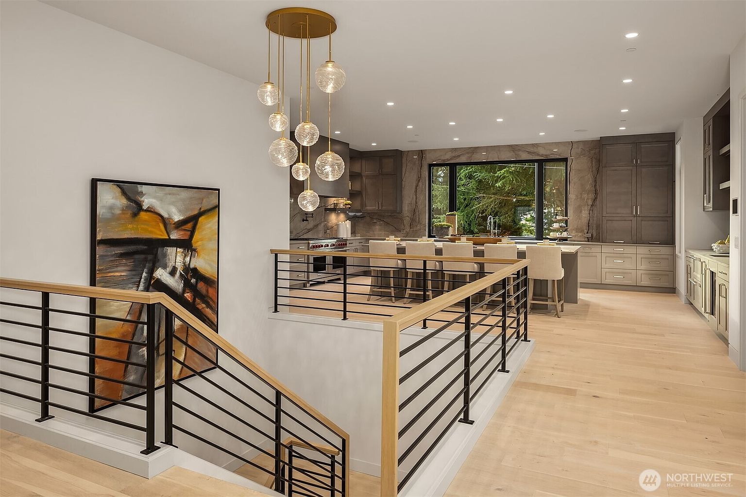 This open-concept kitchen is viewed from an elevated landing, showcasing a large central island with bar seating and a sophisticated multi-globe pendant light fixture. The space features dark cabinetry, a stone-clad wall behind the sink, and light wood flooring that flows throughout the area. A large window above the sink provides a view of the outdoors, creating a bright and airy atmosphere with a modern, high-end aesthetic.