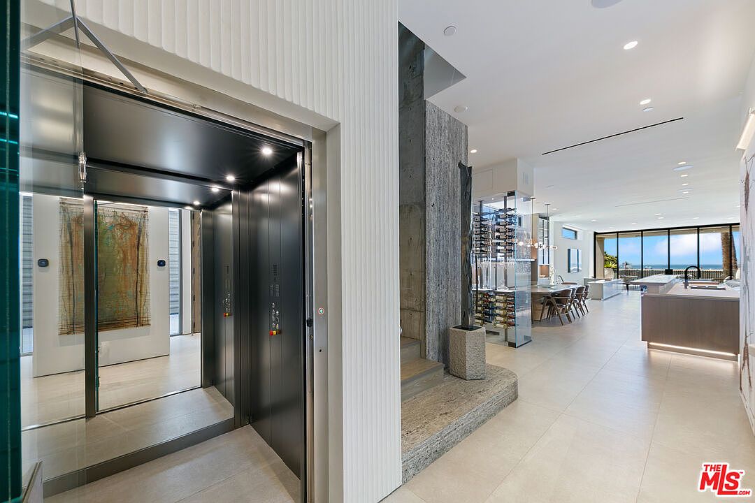 This interior view captures a sophisticated transition space featuring a modern elevator with dark metal doors adjacent to a staircase with a textured stone wall. The perspective looks through the hallway toward an open-concept living and dining area that leads to a bright, expansive view of the ocean. The design aesthetic is minimalist and high-end, characterized by clean lines, neutral tones, and a glass-enclosed wine display.