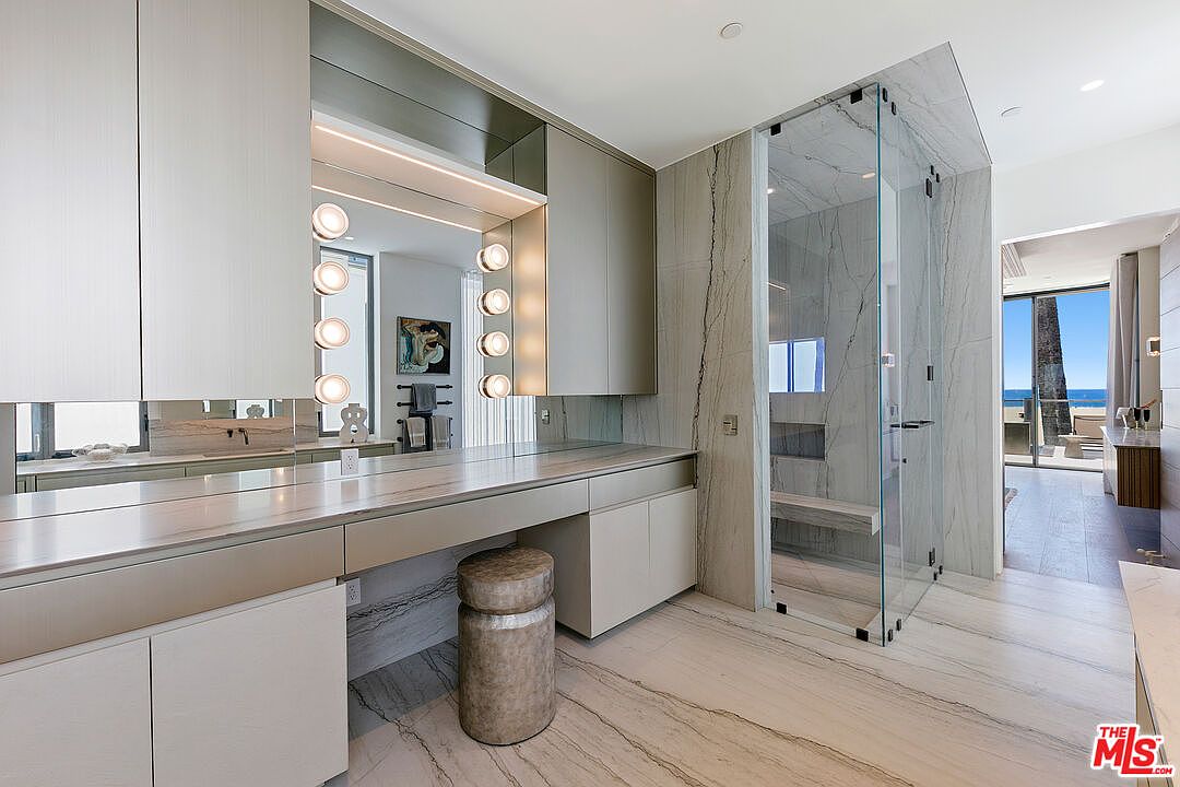 This luxurious primary bathroom features a long, sleek vanity with a stone countertop and integrated lighting, paired with a cylindrical stool. To the right, a glass-enclosed walk-in shower with stone wall cladding offers a clean, modern aesthetic. The space is bright and airy, with a clear line of sight leading through to a sunlit outdoor area with ocean views.