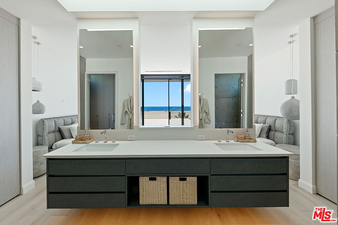 This modern primary bathroom features a dual-sink vanity with a dark, textured finish and a light-colored countertop, centered between two large mirrors. The space is symmetrically designed, with upholstered seating areas on either side and a central window providing a clear view of the beach and ocean. The overall aesthetic is clean, minimalist, and luxurious, emphasizing natural light and a serene coastal atmosphere.