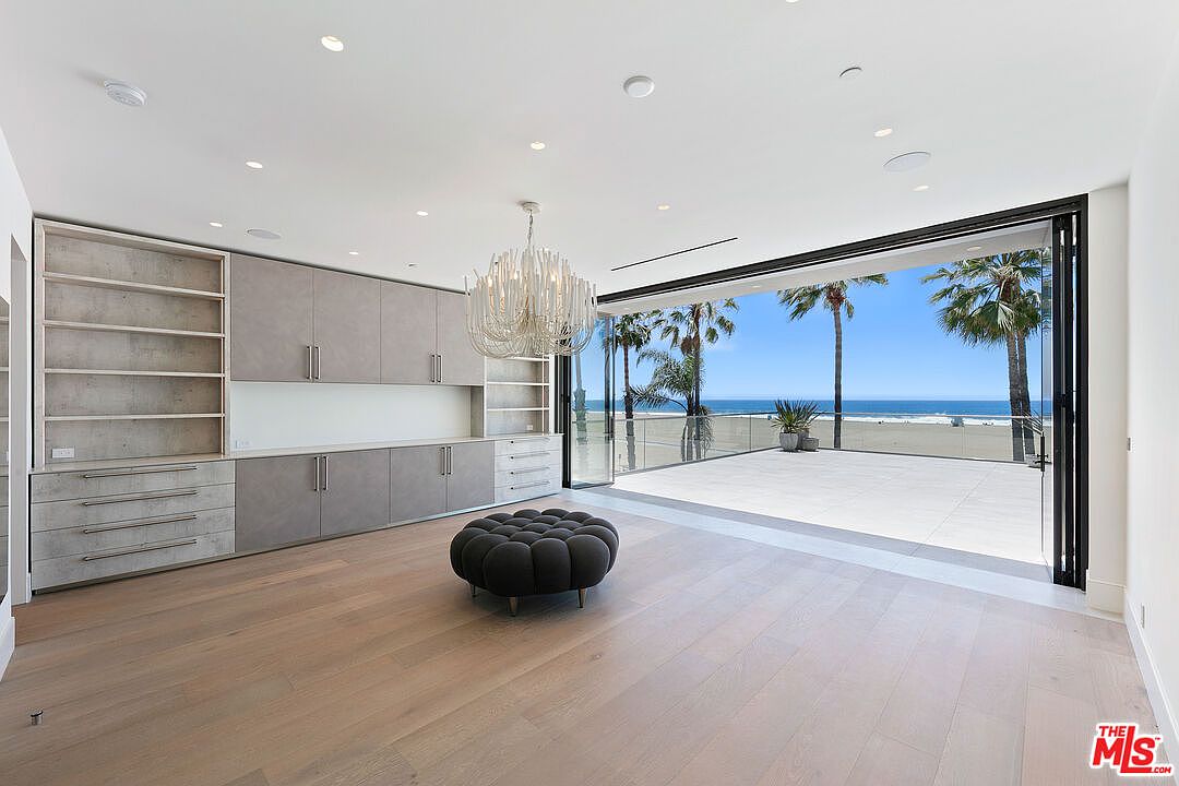 This bright, modern living space features light wood flooring and a large, custom-built storage unit with a mix of open shelving and closed cabinetry. A central, tufted dark gray ottoman sits beneath a unique, sculptural chandelier. The room opens directly onto a spacious balcony through expansive, floor-to-ceiling glass doors that provide an unobstructed, panoramic view of the beach and ocean.