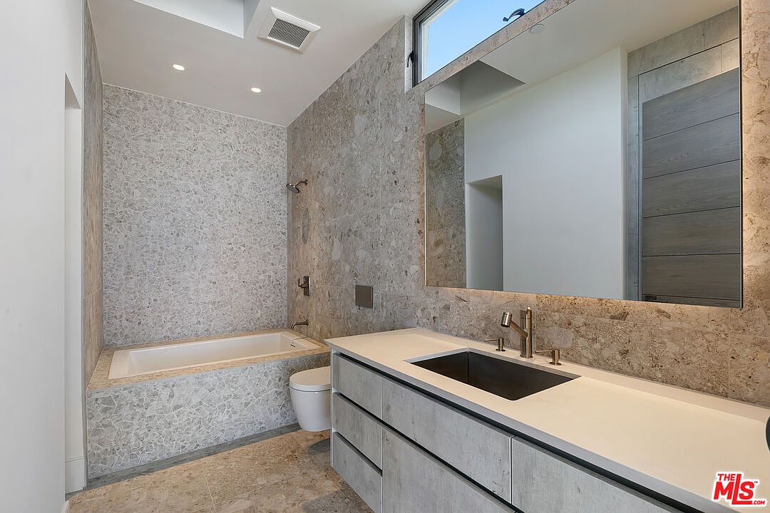 This modern bathroom features a striking stone-clad wall that extends from the vanity area to the built-in bathtub. The space includes a floating vanity with a dark rectangular sink, a large mirror with integrated lighting, and a wall-mounted toilet. A high, horizontal window provides natural light, contributing to the clean, minimalist aesthetic of the room.