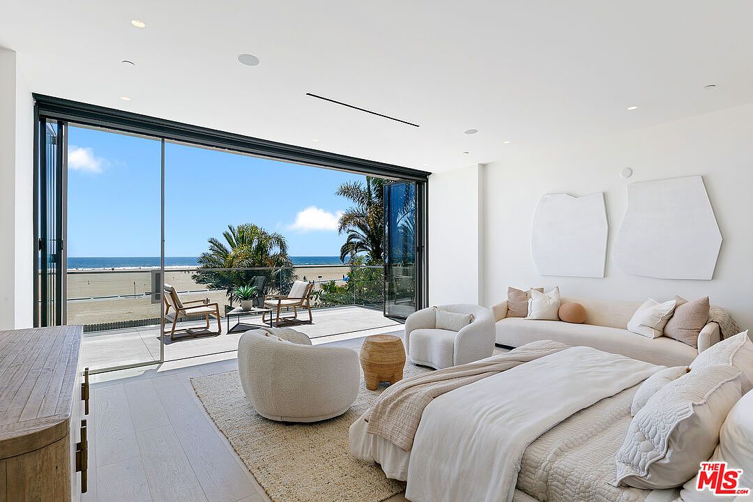 This luxurious primary bedroom features a neutral, minimalist aesthetic with a plush bed, two rounded accent chairs, and a long sofa. The room is defined by expansive floor-to-ceiling glass doors that slide open to reveal a private balcony overlooking the beach and ocean. The bright, airy atmosphere is enhanced by white walls and light wood flooring, creating a serene and sophisticated coastal retreat.