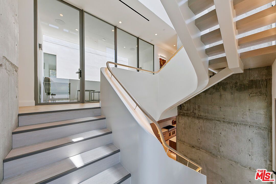 This modern interior features a striking staircase with light wood treads and a sleek, white curved railing that leads to an upper landing. To the left, a glass-enclosed space provides a view into an adjacent room, while a raw concrete wall adds a textured, industrial contrast to the clean lines of the staircase. The perspective captures the architectural flow of the home, highlighting the interplay between natural light and minimalist design elements.