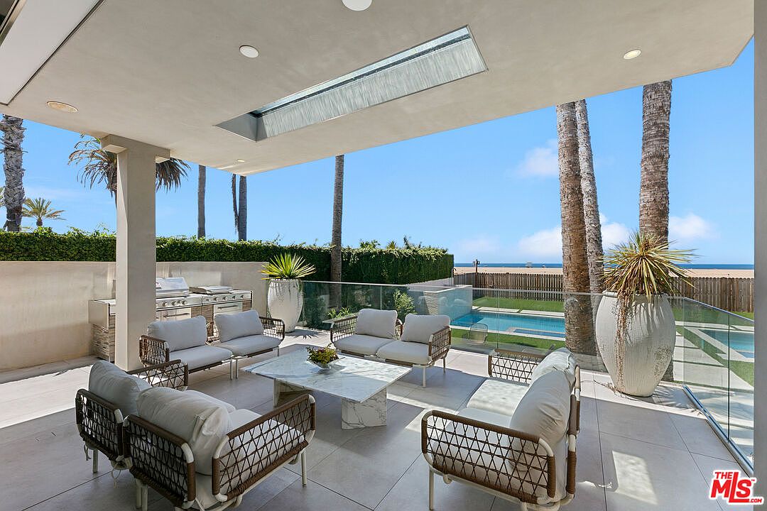 This luxurious covered patio offers a seamless transition between indoor and outdoor living, featuring a comfortable seating area with modern wicker furniture and a central stone-topped coffee table. The space includes an outdoor kitchen setup and overlooks a private swimming pool, with a clear glass railing providing unobstructed views of the beach and ocean beyond. The setting is bright and airy, emphasizing a sophisticated coastal lifestyle.