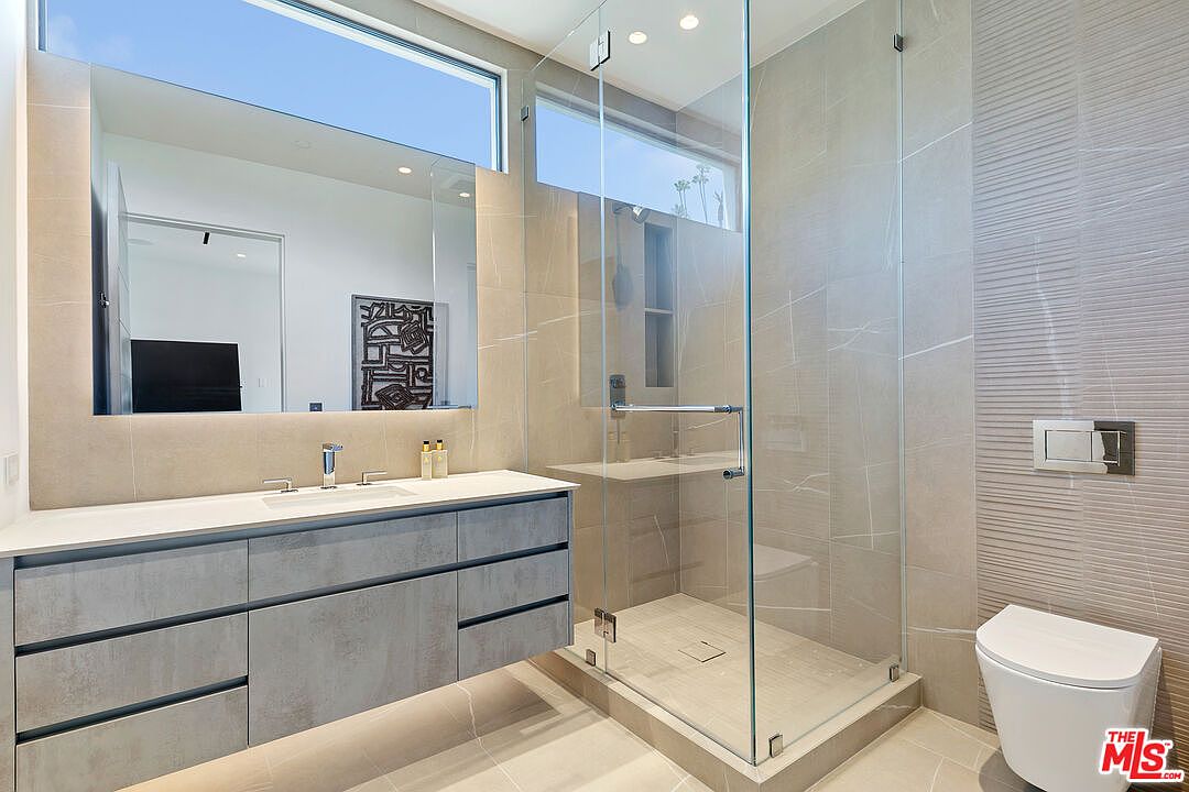 This modern bathroom features a floating vanity with a stone countertop and a large, rectangular mirror positioned beneath a horizontal window. To the right, a glass-enclosed walk-in shower with neutral-toned wall tiles sits adjacent to a wall-mounted toilet. The space is characterized by a clean, minimalist aesthetic with a neutral color palette and contemporary fixtures.